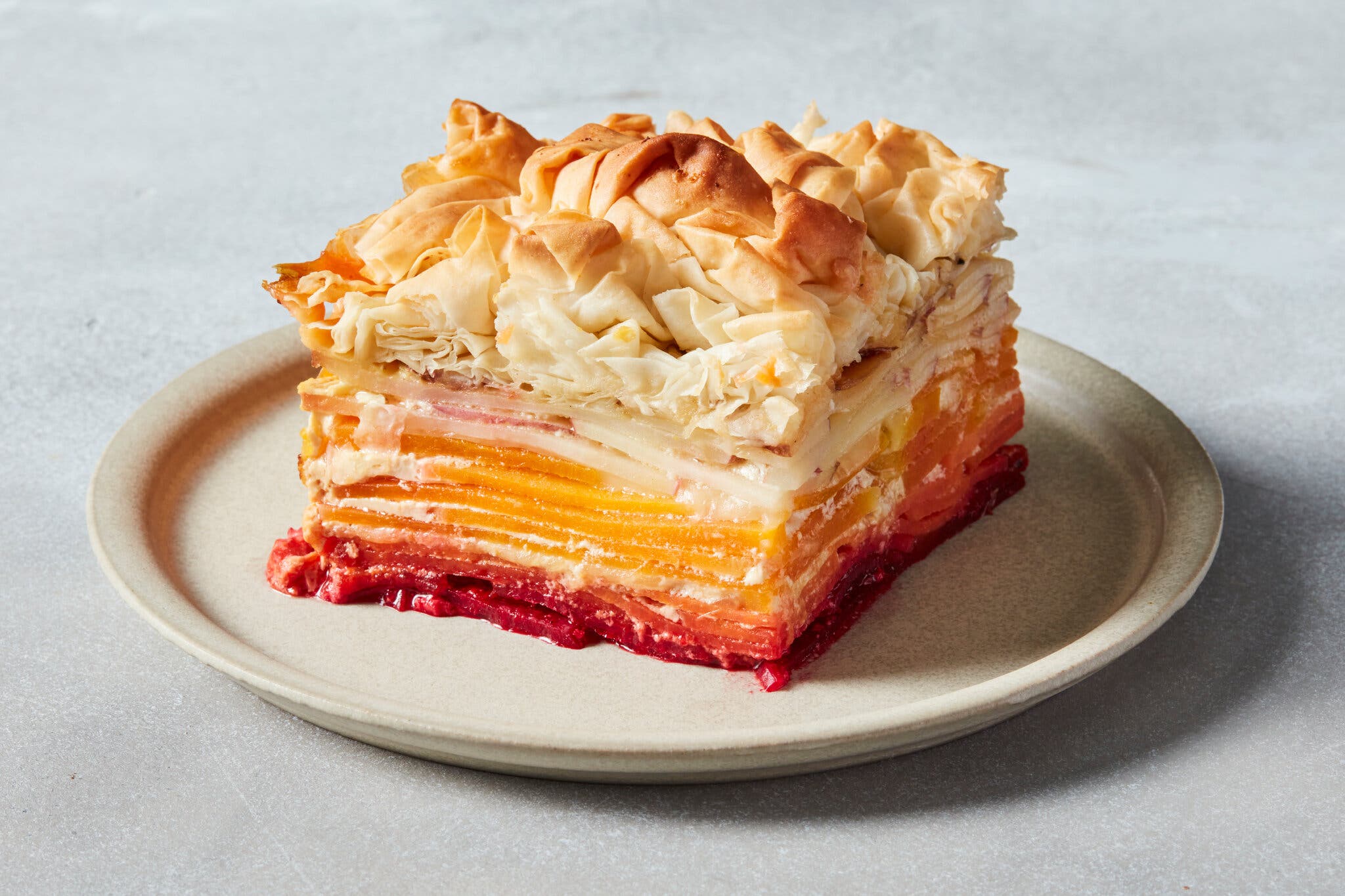 A square slice of a casserole layered with thinly sliced red, orange and white vegetables from darkest at the bottom to lightest at the top. The whole thing is topped with crinkled phyllo dough.