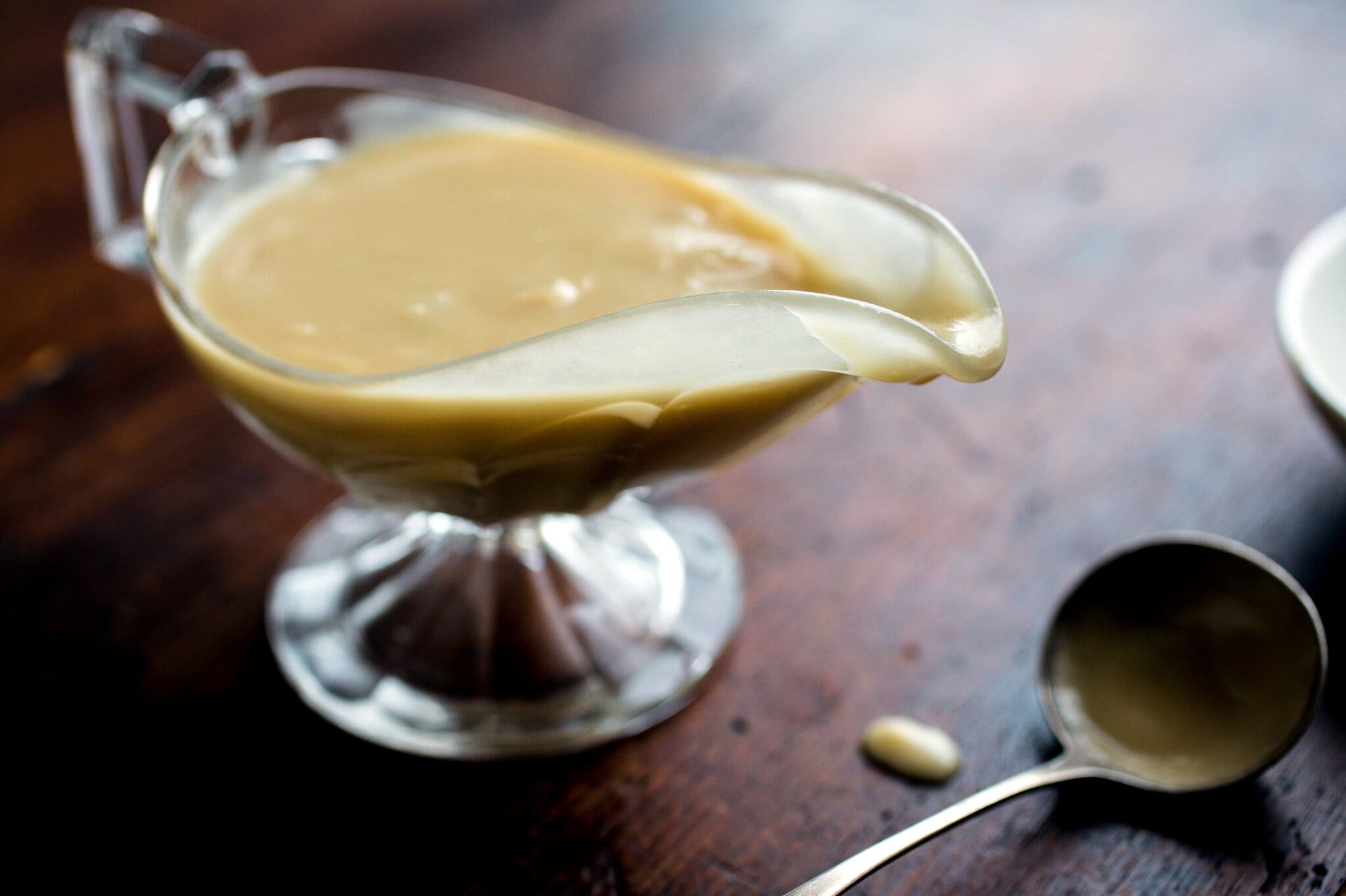 Light brown gravy fills a clear glass gravy boat on a wooden surface. A silver ladle sits nearby.