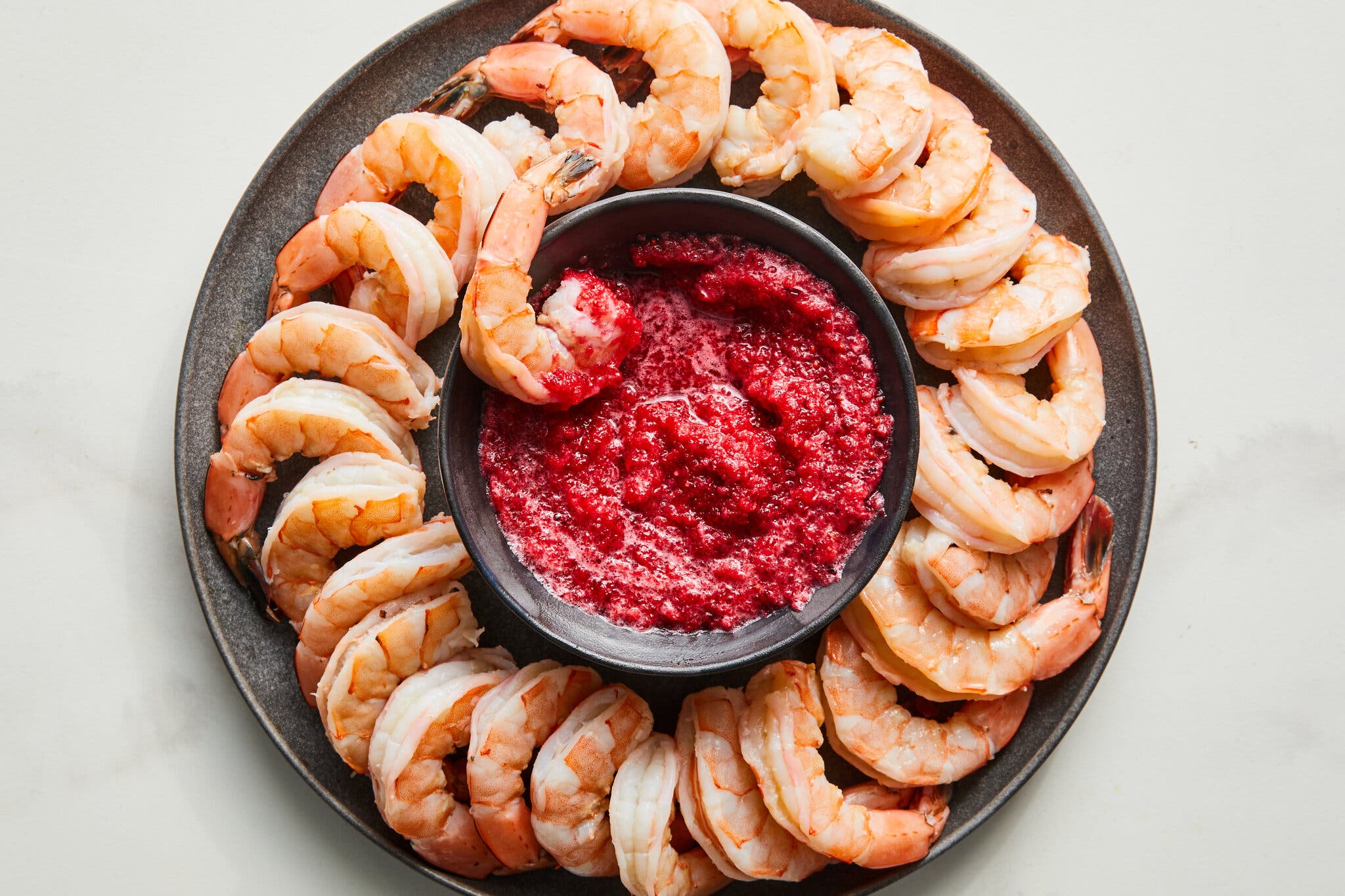 Cooked shrimp arranged in a circle on a dark gray platter, surrounding a small bowl of red sauce. One shrimp is dipped into the sauce.