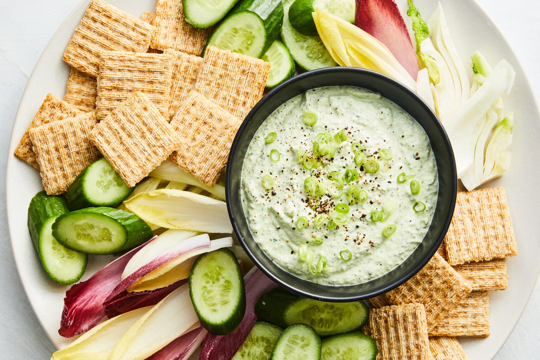 A round white platter holds a black bowl of creamy green dip garnished with chopped green onions. Around it are square crackers, cucumber slices and endive leaves.