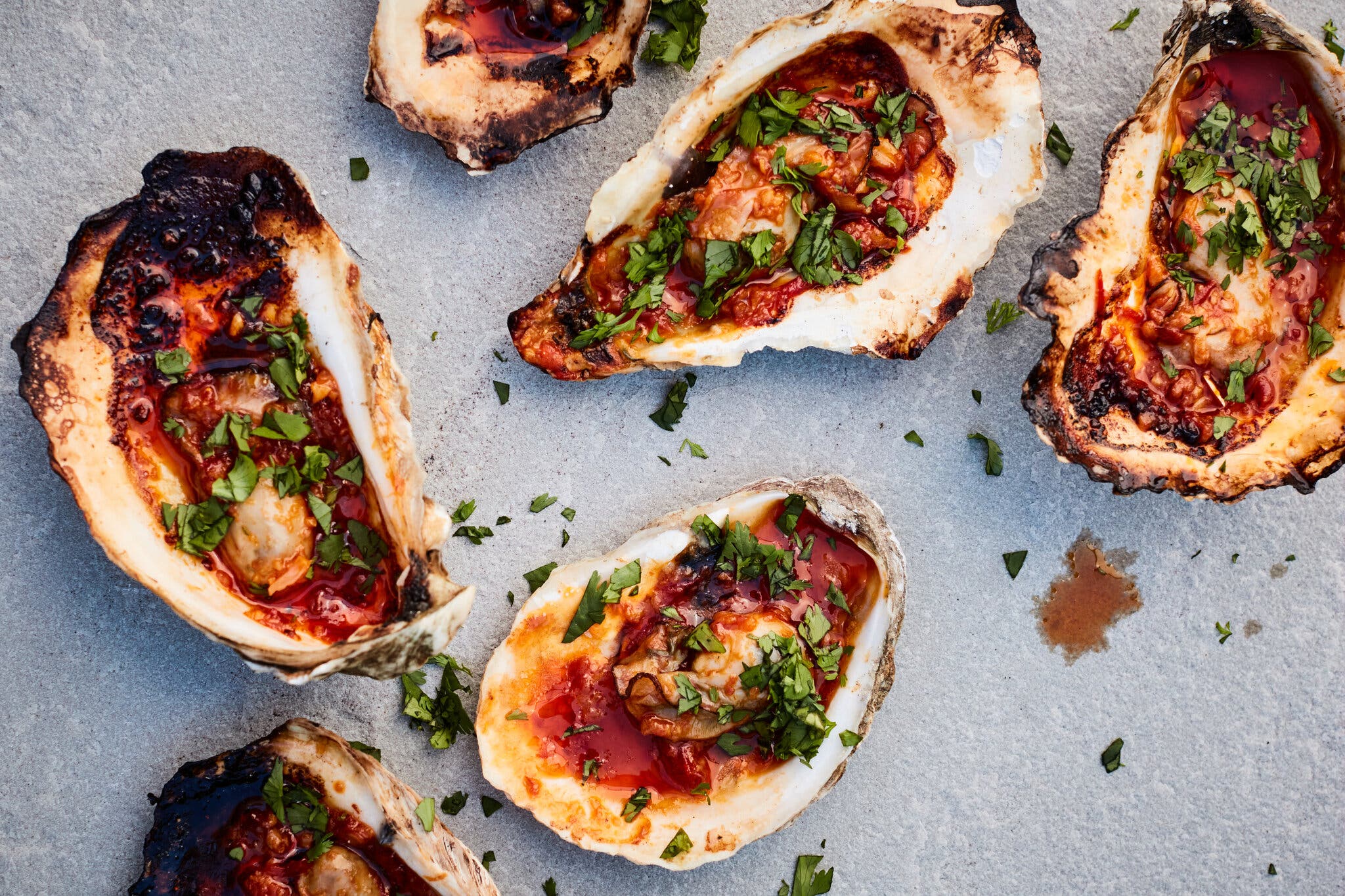 Oysters, some of which are a bit charred, with red sauce and green herbs scattered on top.