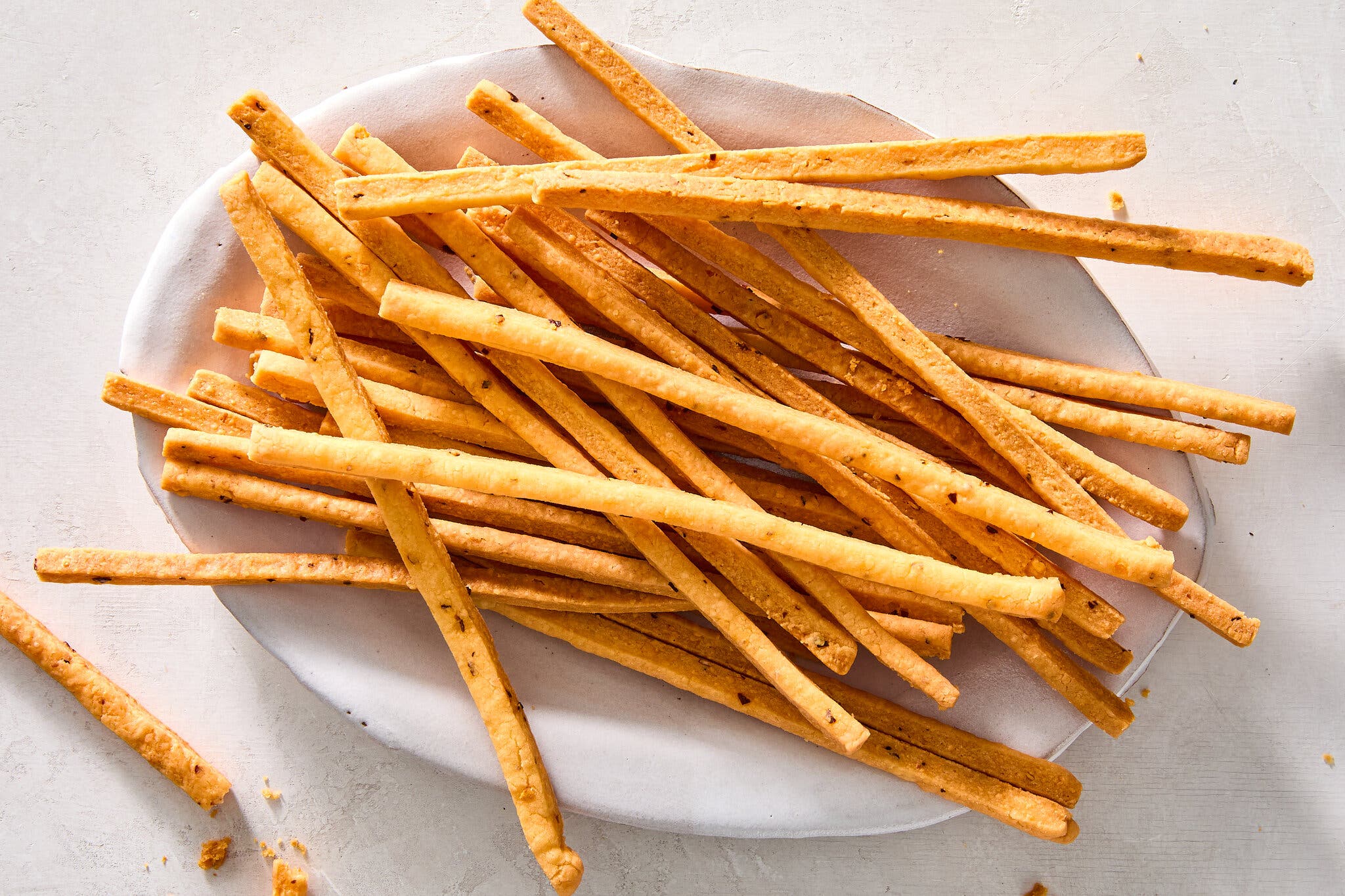 A white plate is piled high with golden-brown, thin breadsticks.