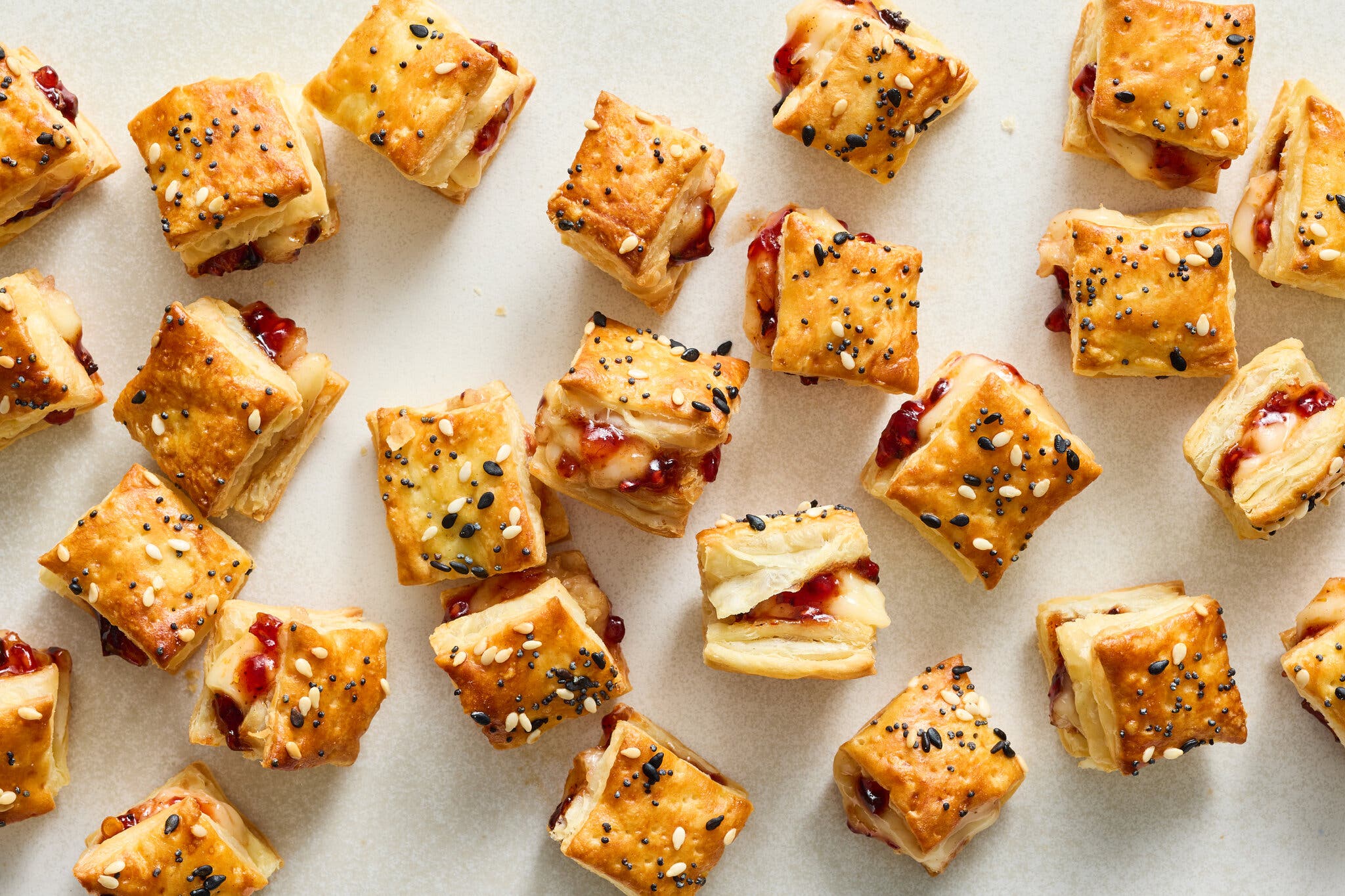 Golden-brown, square pastries filled with red jam and white, creamy cheese are arranged haphazardly. Each pastry is sprinkled with black and white seeds.