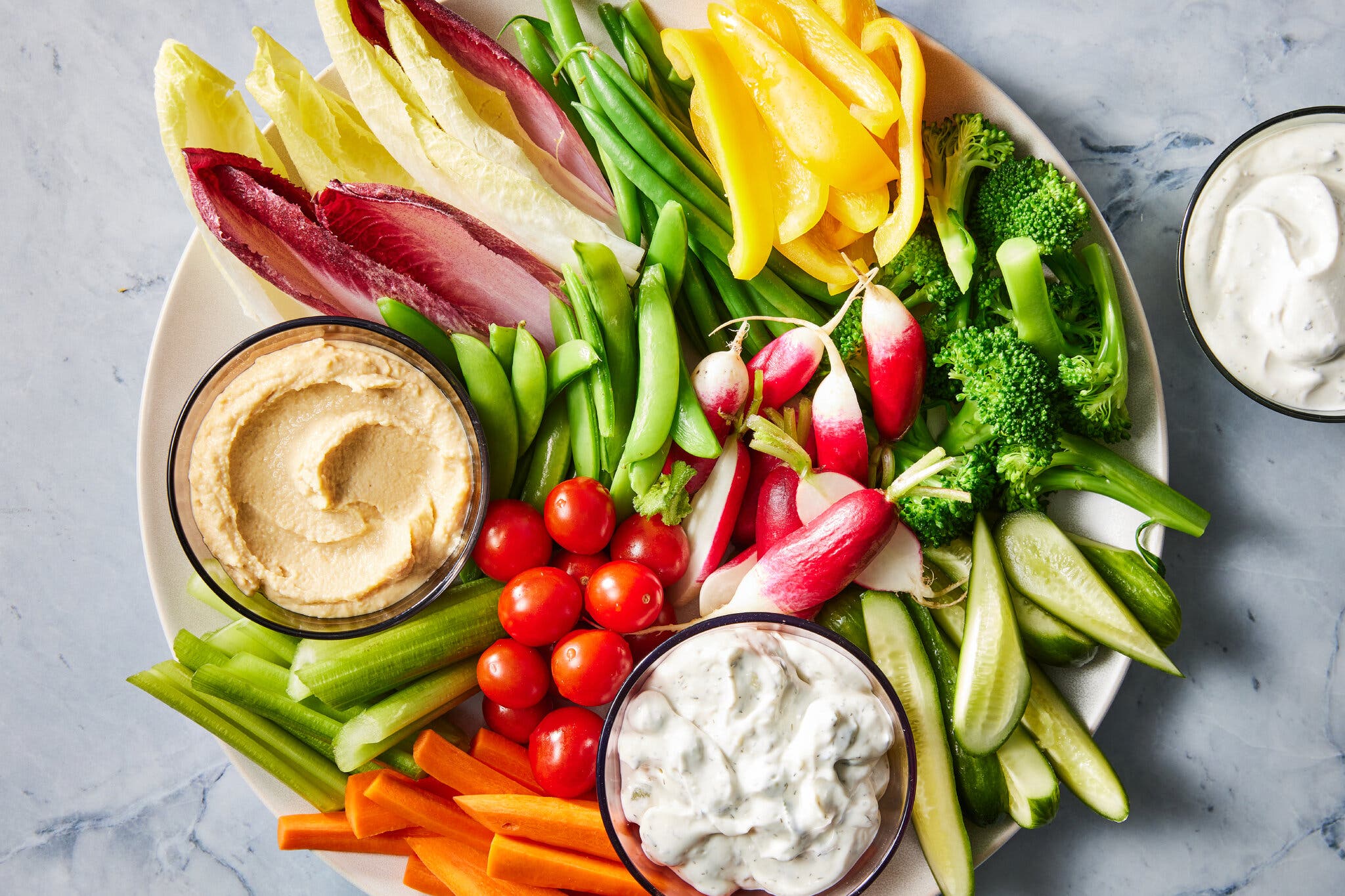 A white platter filled with a colorful array of raw and blanched vegetables, including yellow peppers, green beans, radishes, carrots and celery. Three small bowls of dip, one hummus and two creamy white, are arranged among the vegetables.