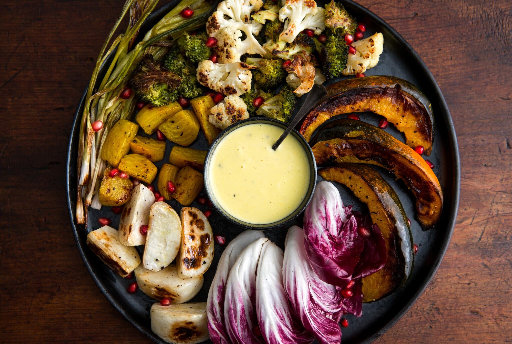 Various roasted vegetables are arranged on a black platter, with a small bowl of yellow sauce in the center.