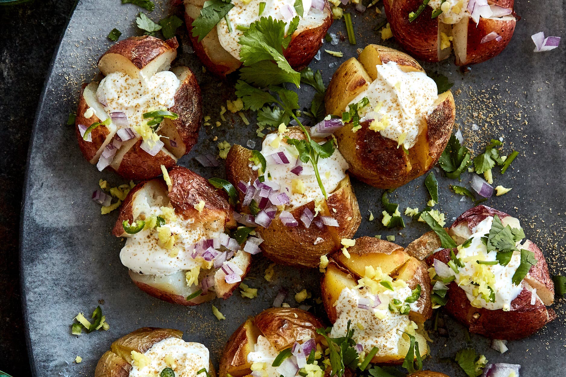 Small roasted potatoes, split and topped with cream, red onions, green herbs and yellow zest, on a dark surface.