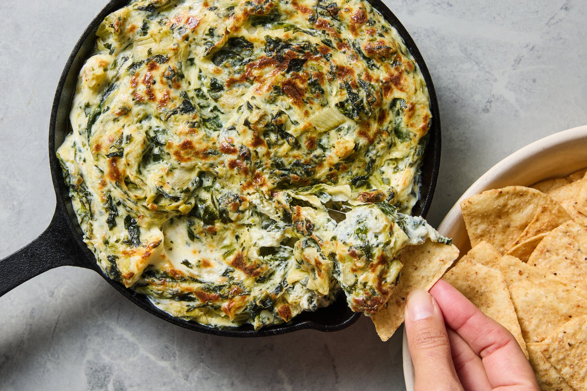 An overhead view of a hand dipping a triangular tortilla chip into bubbling, cheesy spinach and artichoke dip in a black cast-iron skillet.