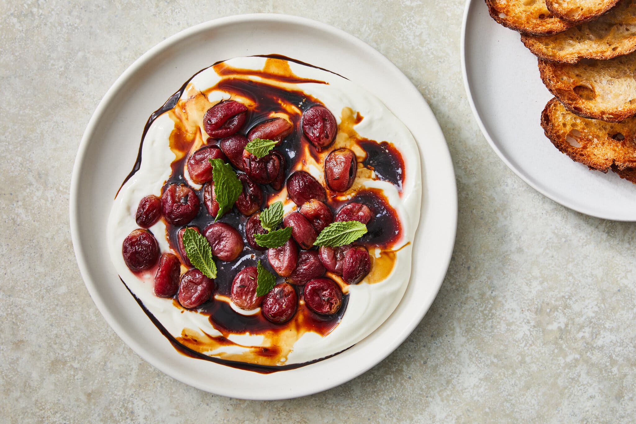 A white plate filled with creamy white dip topped with roasted grapes, a brown drizzle and fresh mint leaves. Another plate of tasted bread sits nearby.