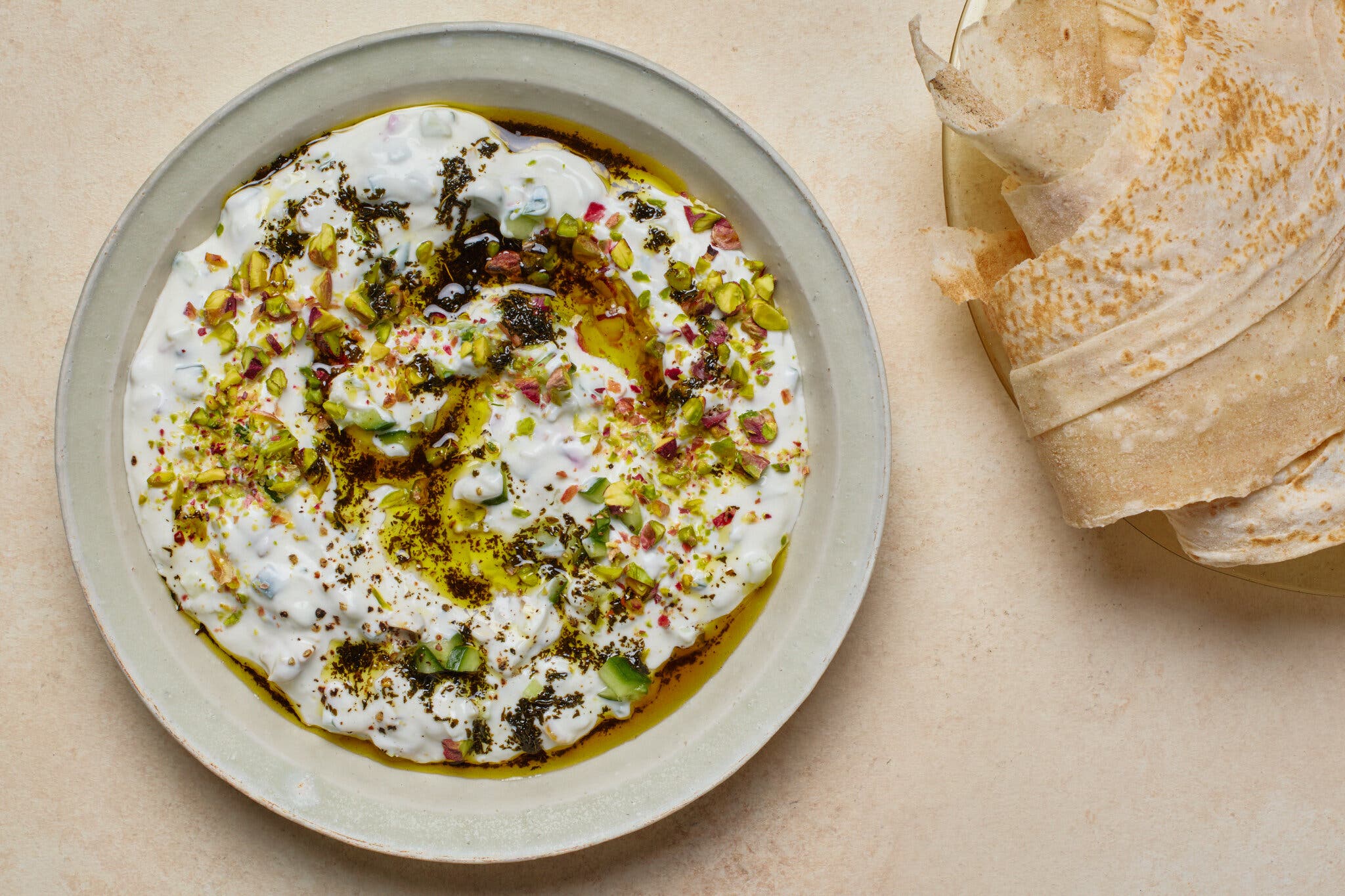 A creamy white dip topped with chopped pistachios, herbs and olive oil. Stacked flatbreads rest beside the plate.