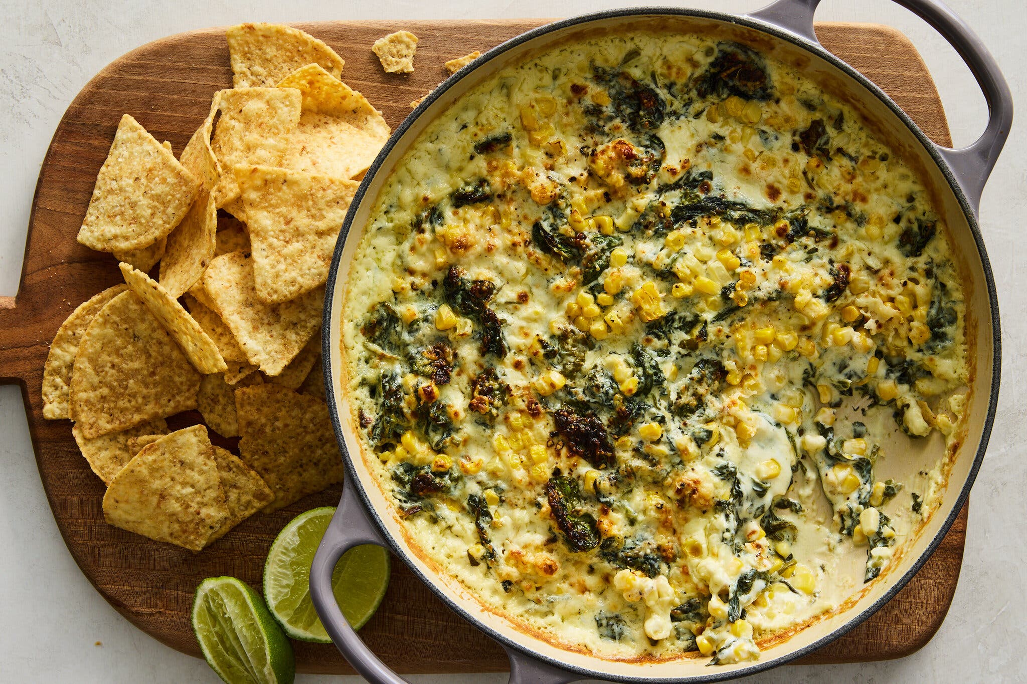 A large, creamy skillet dip featuring yellow corn kernels and wilted dark green spinach, baked until golden brown on top. The dip is surrounded by light brown tortilla chips and two bright green lime halves, all resting on a round wooden cutting board.