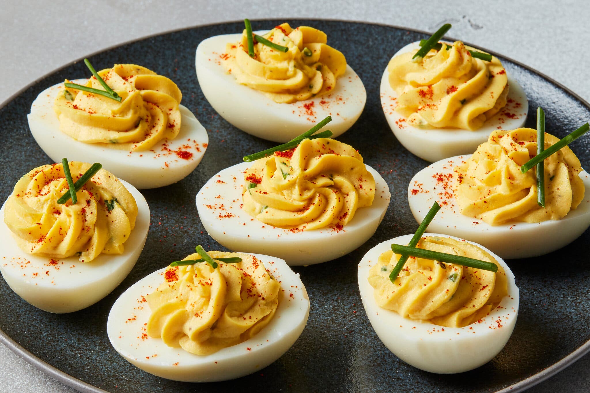 Eight deviled eggs arranged on a dark plate, each filled with swirled yellow yolk, sprinkled with red paprika and topped with green chives.