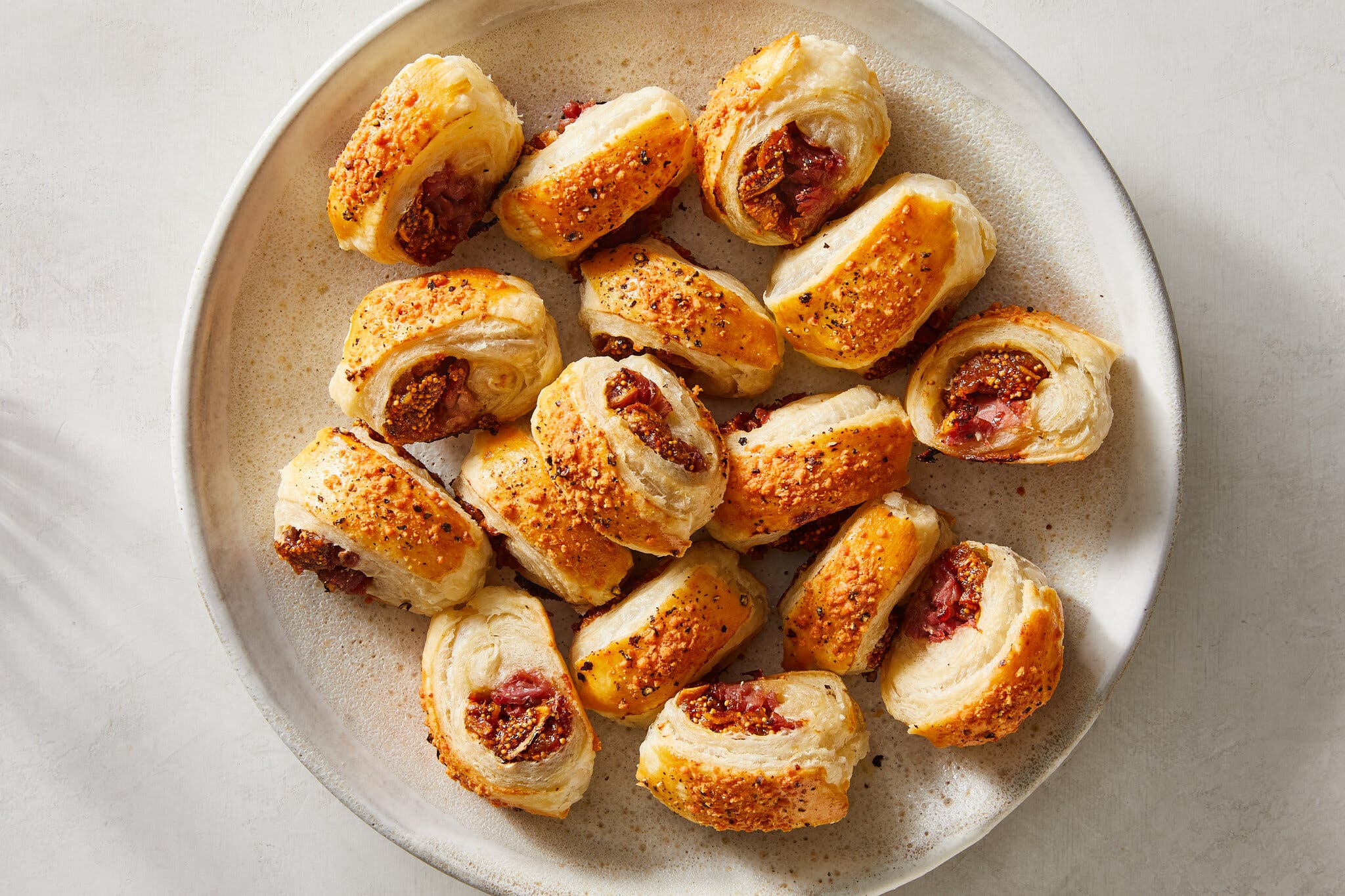 Many golden-brown pastry rolls, with figs and meat visible in their centers, arranged on a light gray plate.