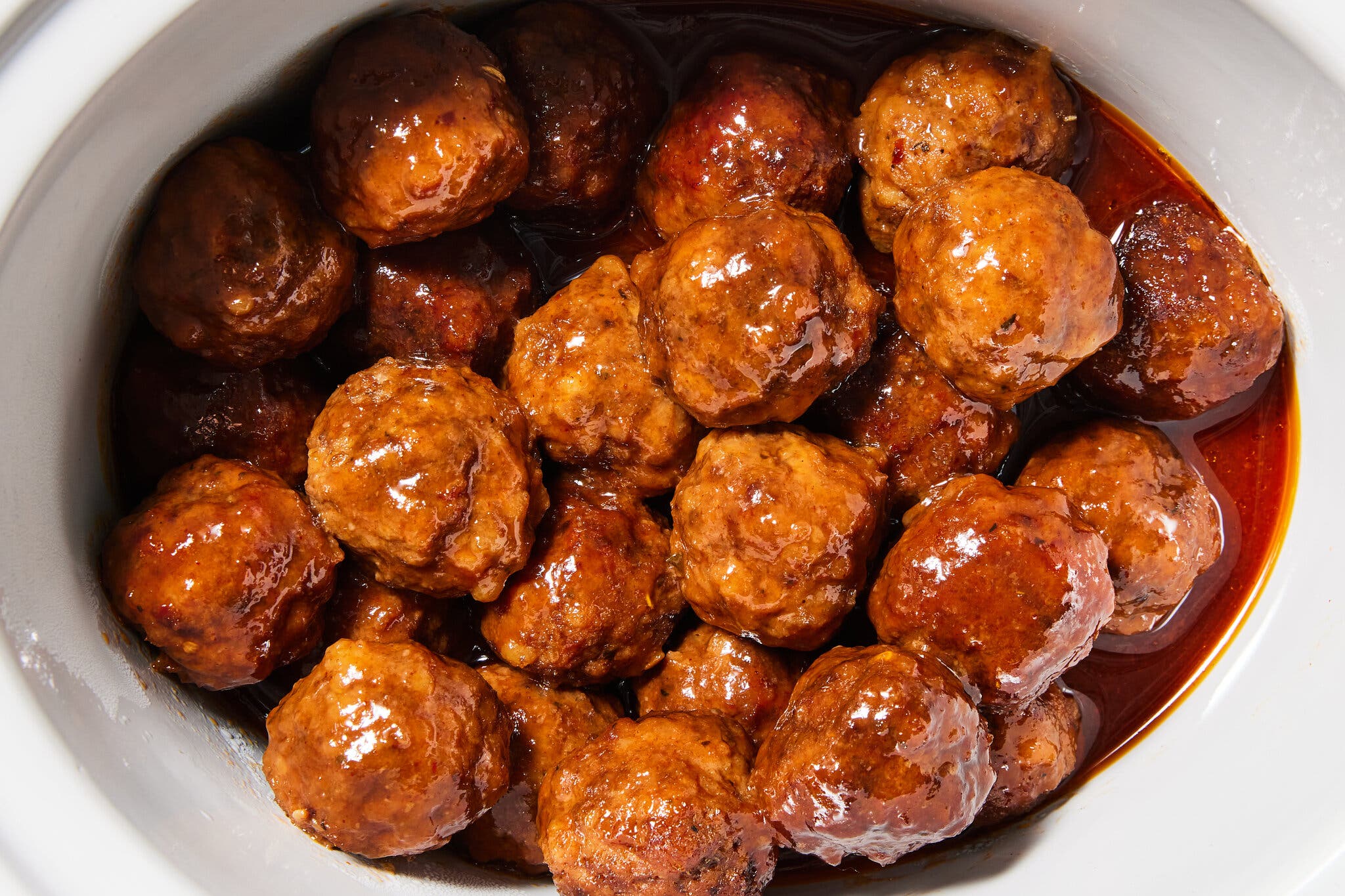 Round, brown meatballs glisten with a thick, dark sauce inside a white cooking vessel.