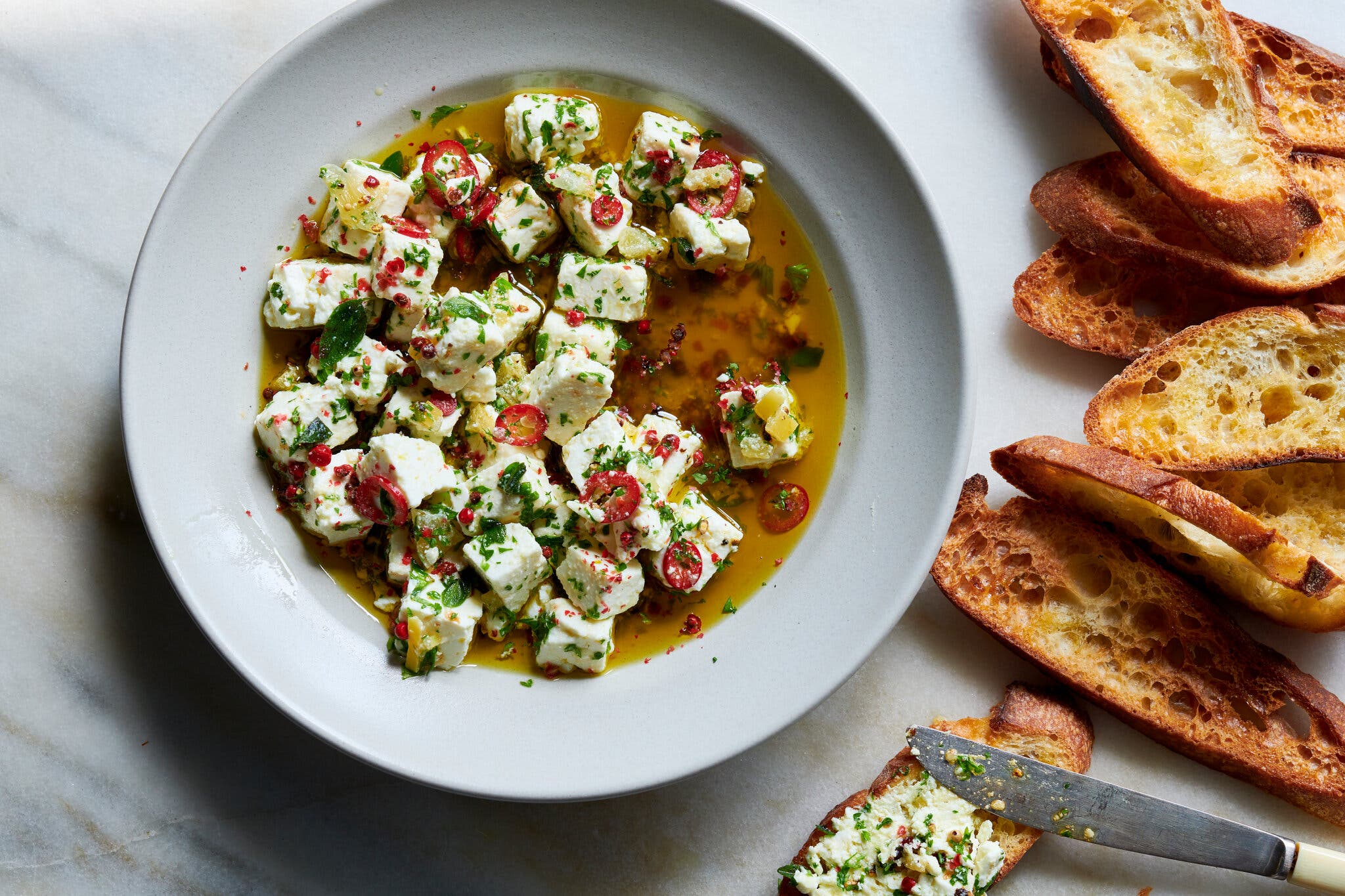 A white bowl holds cubed cheese in yellow oil, topped with green herbs and red peppers. Toasted baguette slices are next to it.