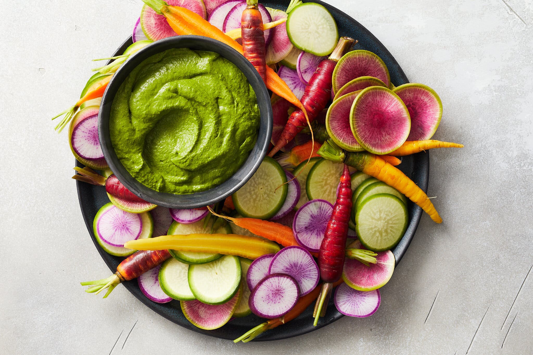 A dark bowl of green dip sits on a plate. Colorful carrots and sliced pink, purple and green radishes surround it.