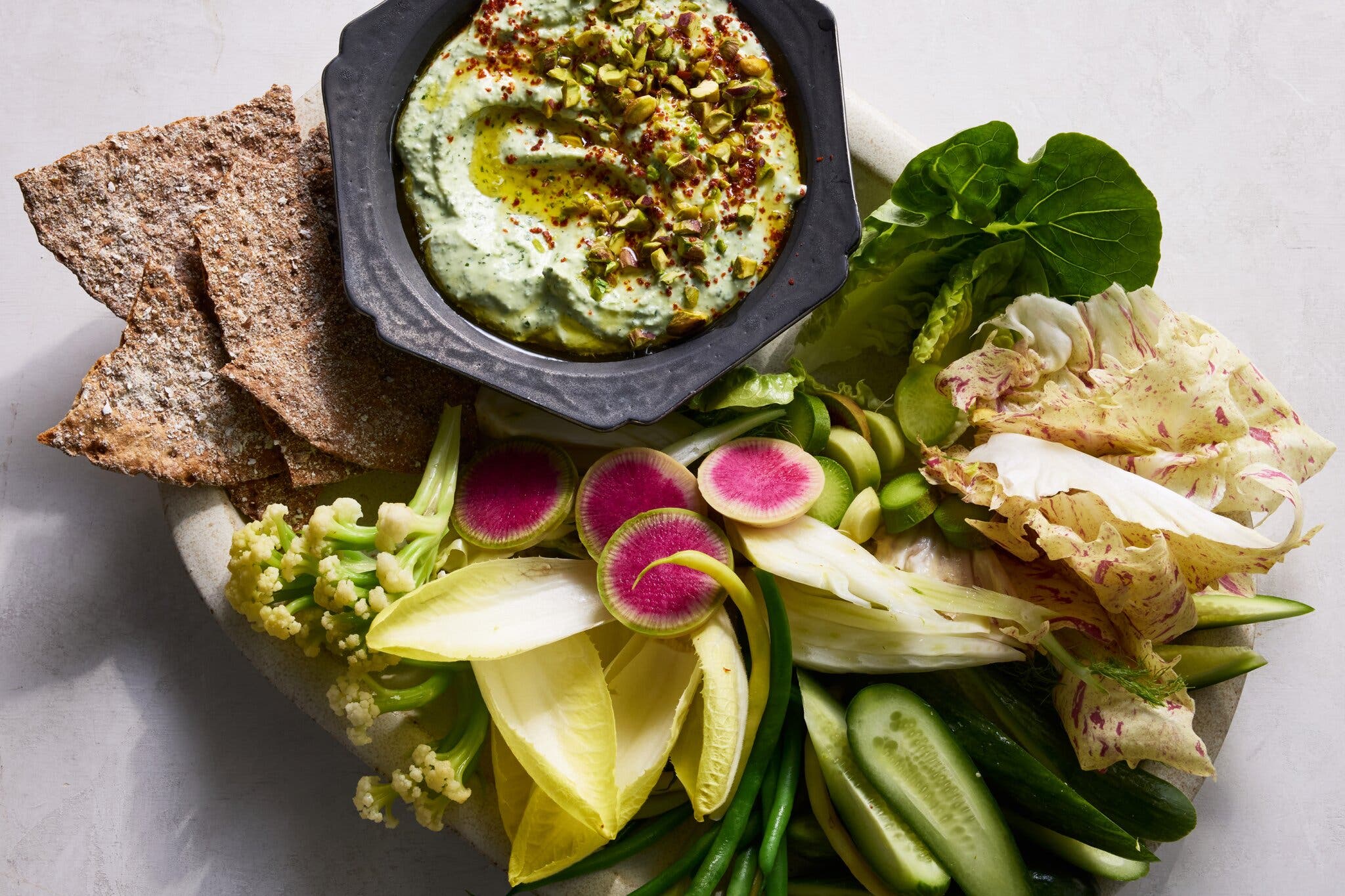 A platter of raw vegetables like sliced cucumber and radish, arranged around a bowl of pale green dip topped with pistachios and oil.