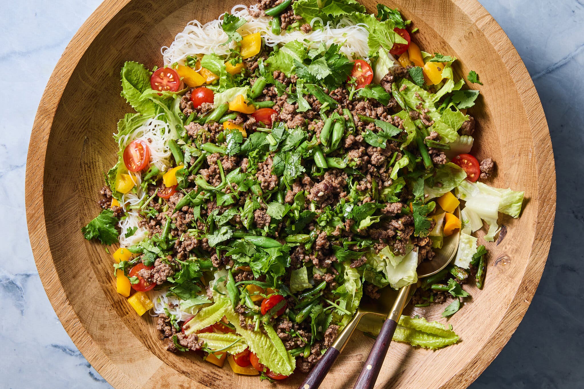 A close-up, overhead view of a vibrant salad with ground beef, green beans, yellow peppers and cherry tomatoes served over white rice noodles in a large wooden bowl.