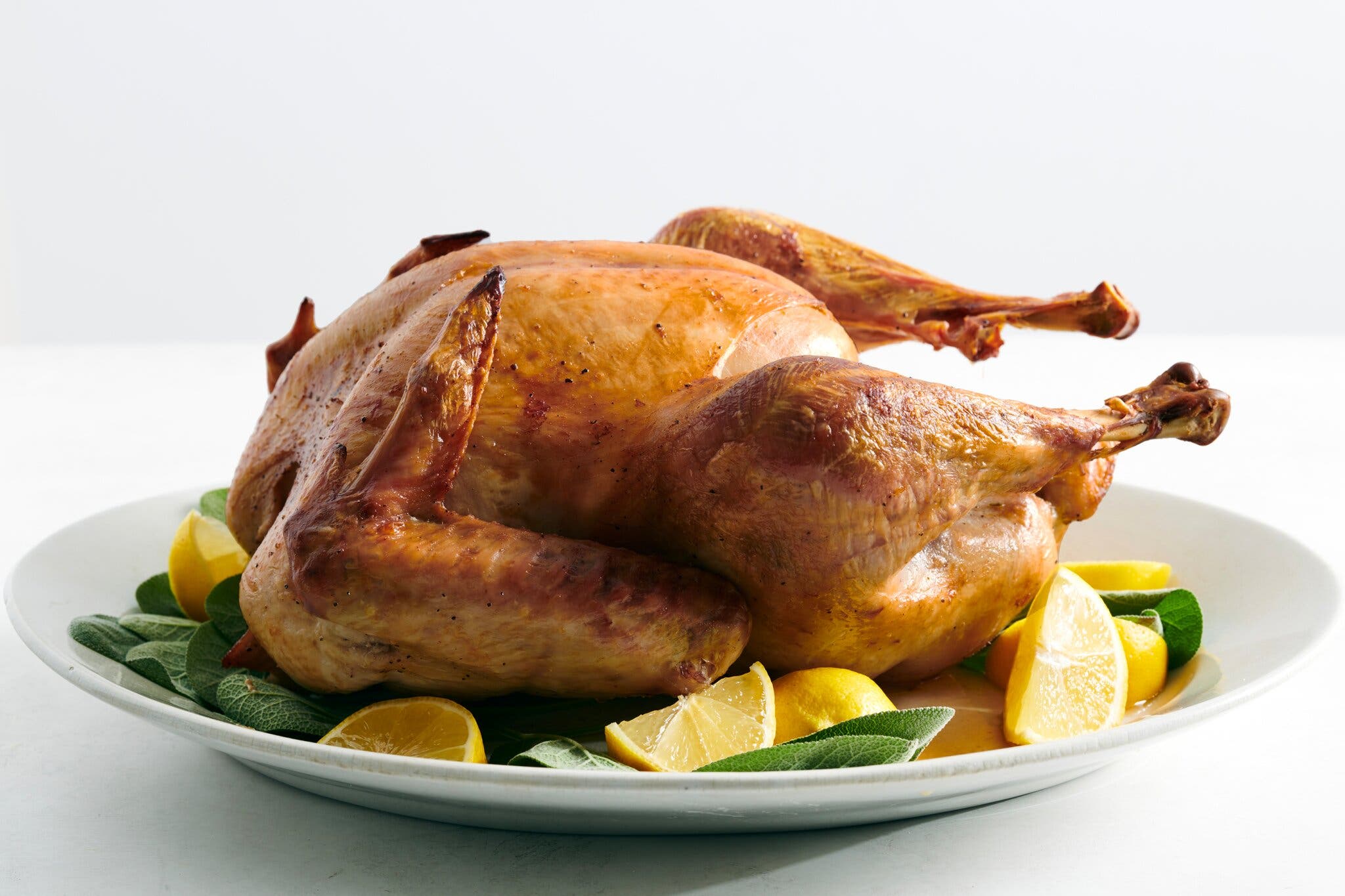 A golden-brown roasted turkey sits on a white platter. It is surrounded by bright yellow lemon wedges and green sage leaves.