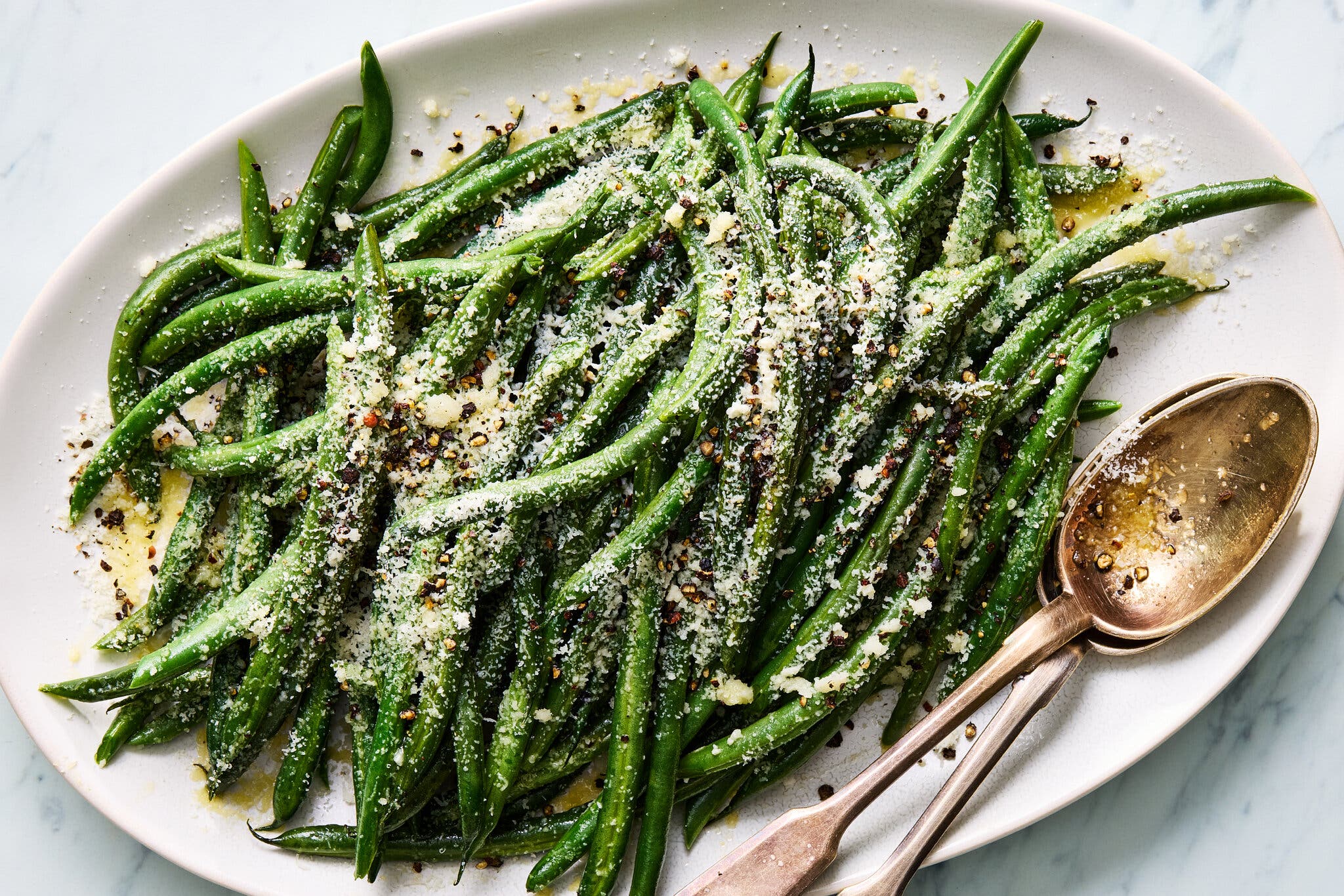 An overhead shot of a white plate of cacio e pepe green beans. Two gold metal spoons sit at the bottom right corner.