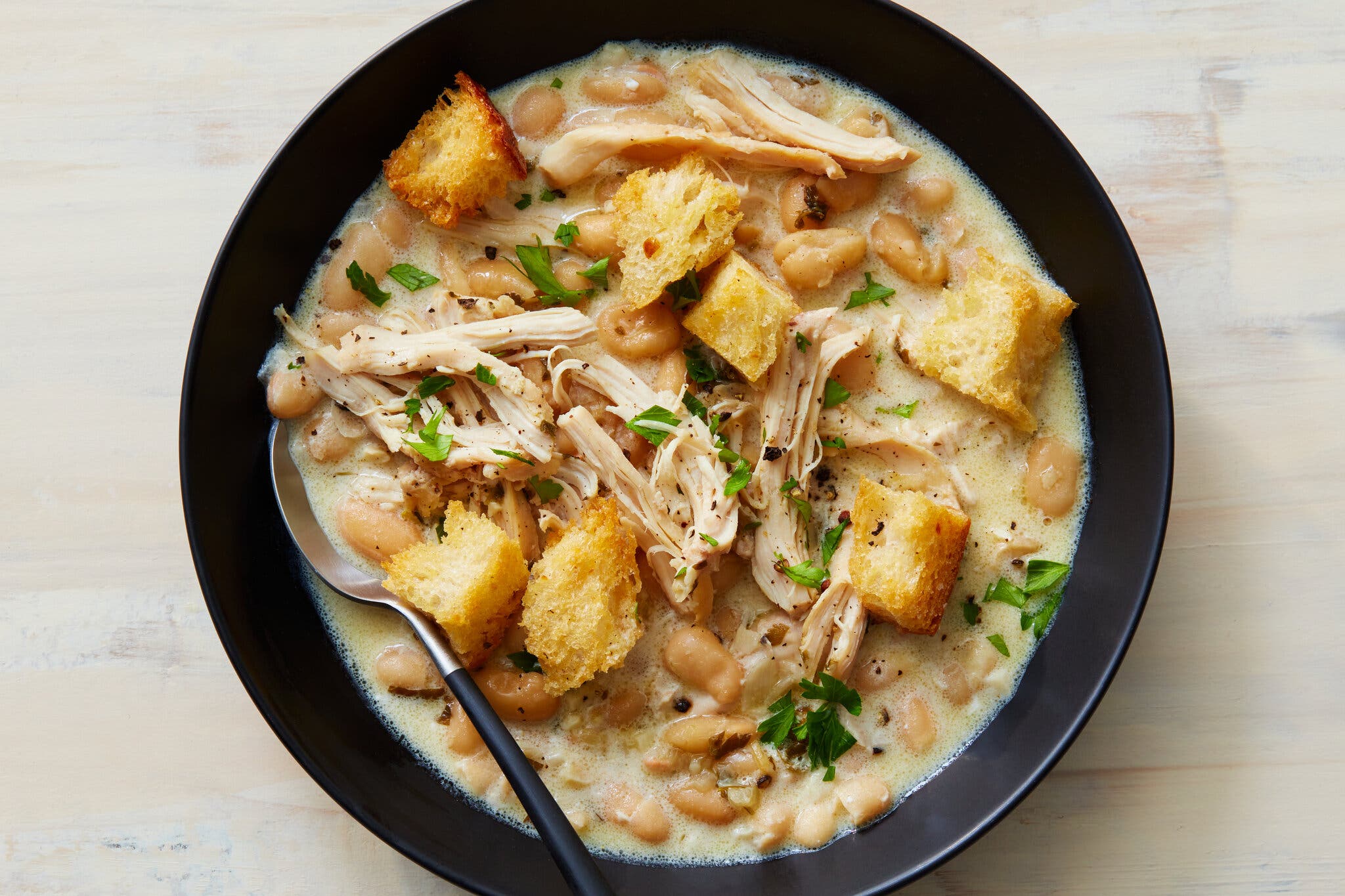 An overhead image of a black bowl filled with braised chicken and beans topped with croutons.