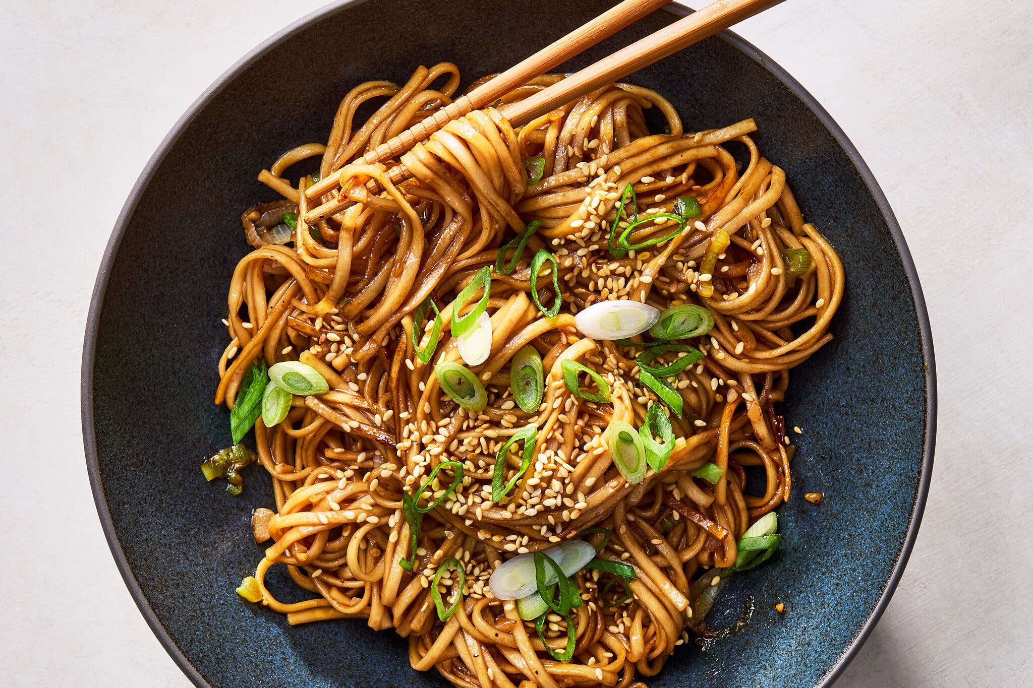 A dark blue bowl filled with noodles in a glossy brown sauce, garnished with sesame seeds and sliced green onions, with chopsticks resting on top.