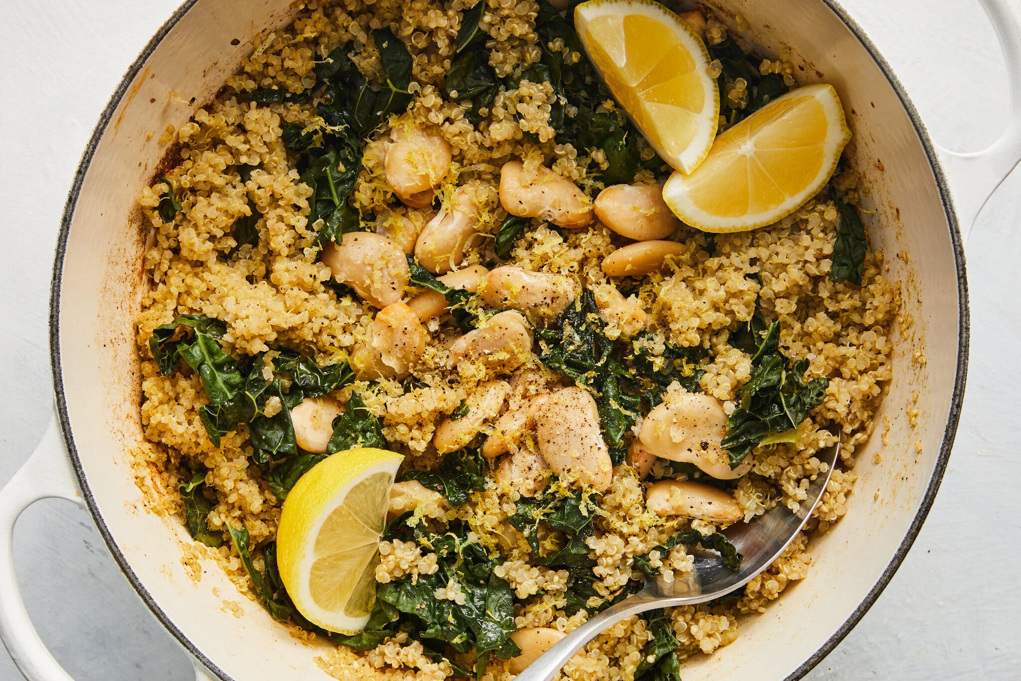 A white pot filled with quinoa, dark green kale, and large white beans, topped with lemon zest and three lemon wedges.