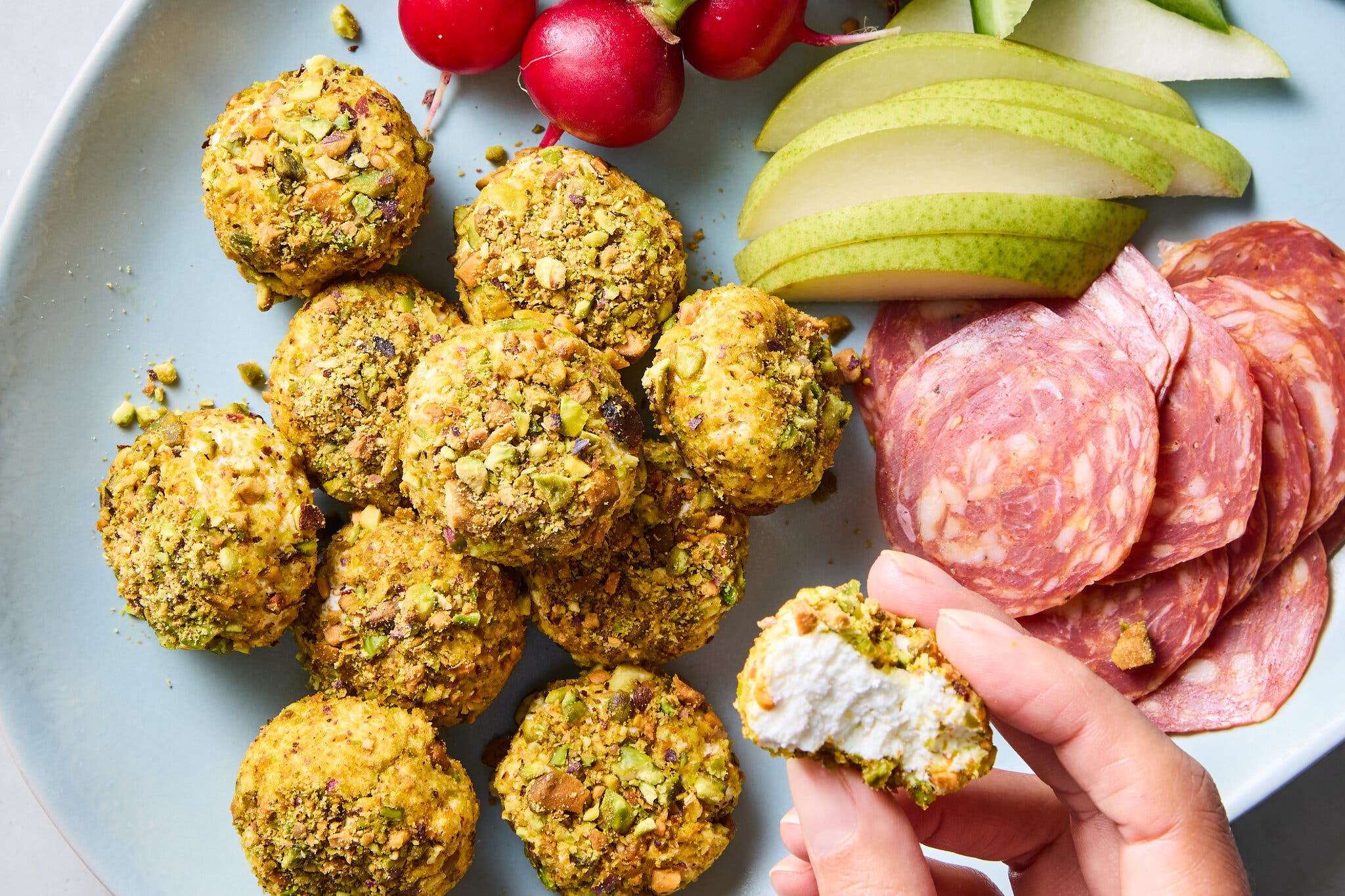 A charcuterie plate with pistachio-crusted goat cheese balls, slices of salami, apple slices, cucumber spears and small radishes.