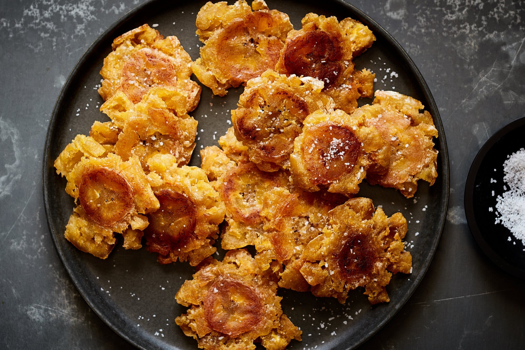 A black plate of tostones topped with salt. A small dish of salt lies to the right.