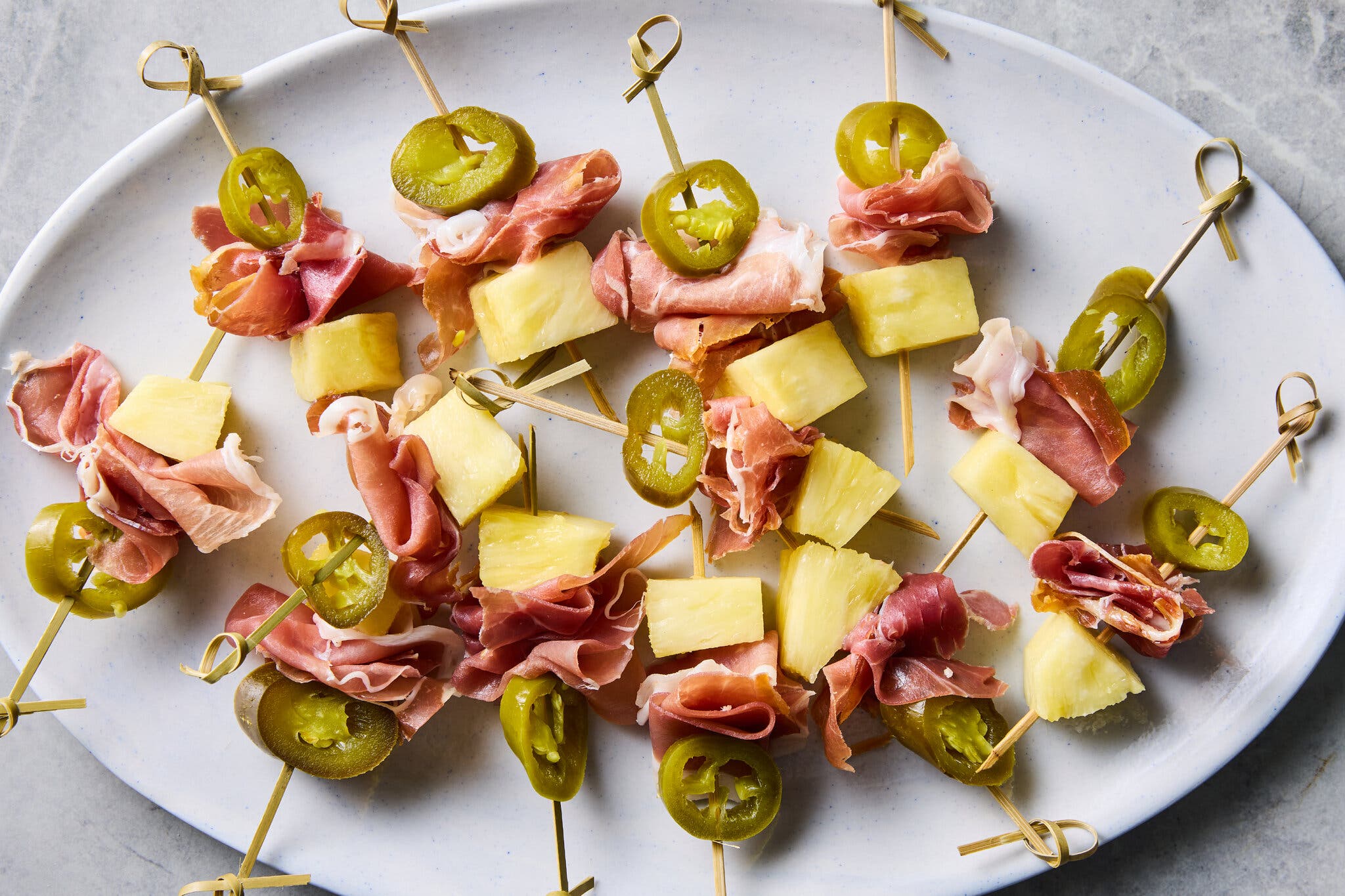 An oval platter with skewers of pineapple, prosciutto and pickled jalapeño slices.