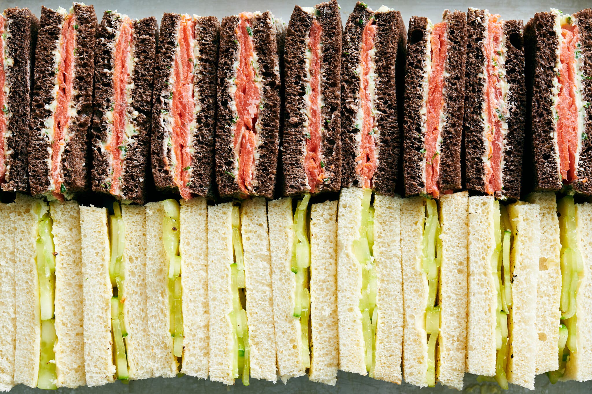 Two rows of tea sandwiches lie cross-section up. The top row is smoked salmon on pumpernickel bread, and the bottom is cucumber on white bread.