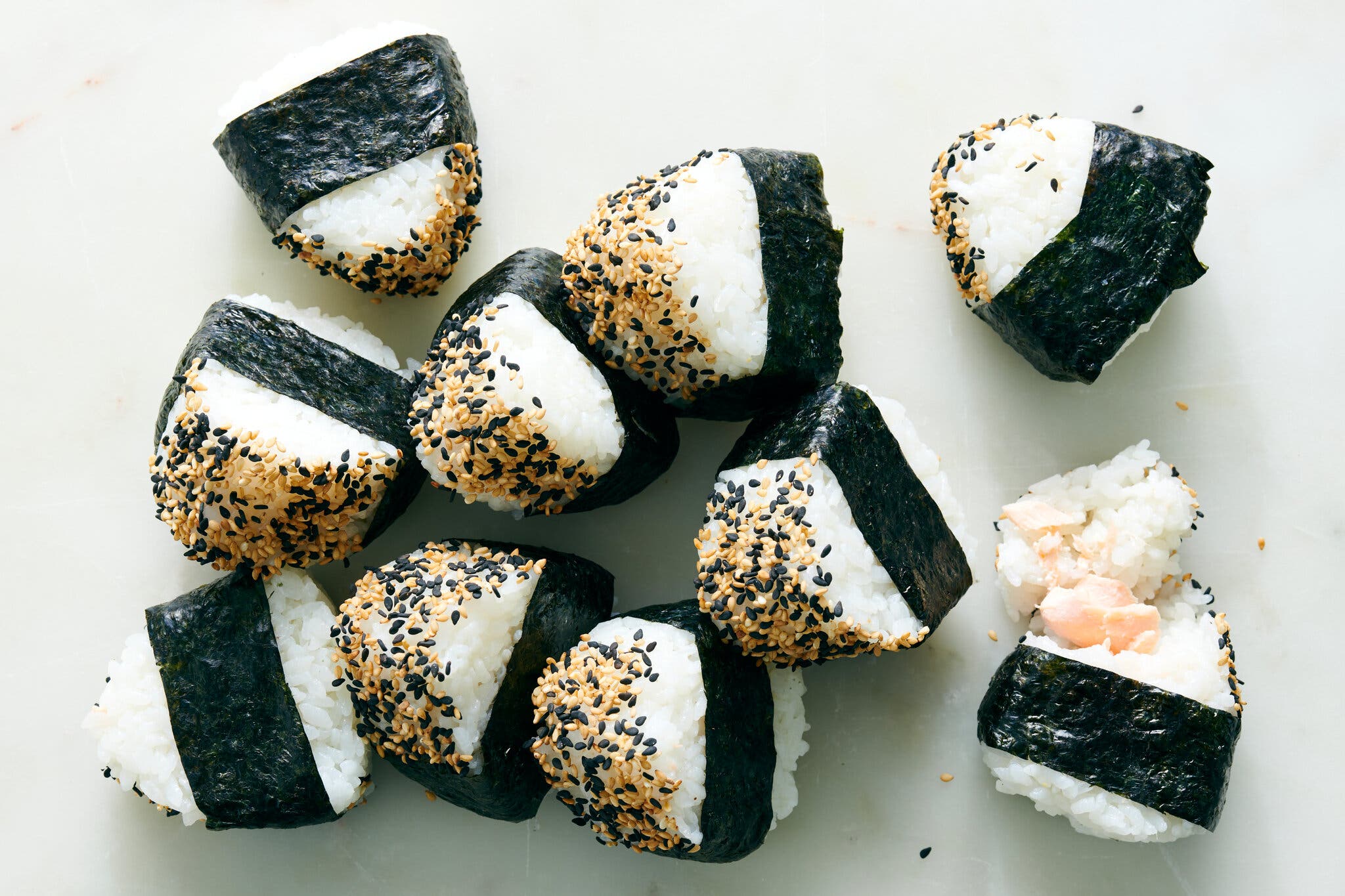 Onigiri with black and white sesame seeds sprinkled on the top. In the bottom right, one rice ball is broken open to show flaked salmon inside.