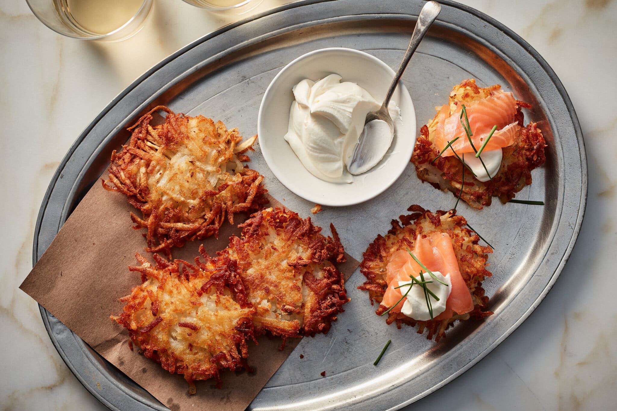 A platter of potato latkes, some topped with chives, sour cream and smoked salmon. There is a small bowl of sour cream with a spoon in it.