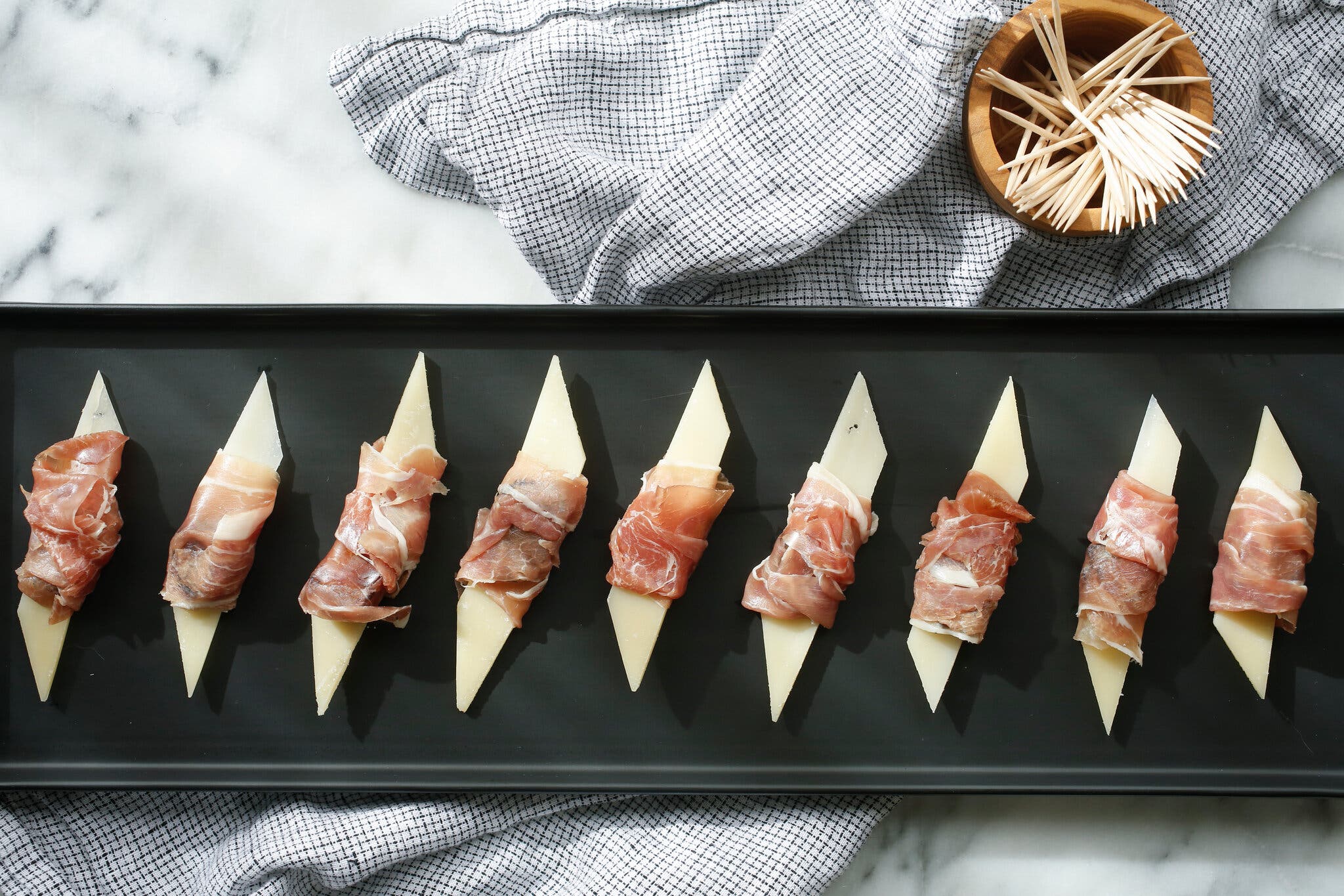 A row of slices of Parmesan cheese wrapped with prosciutto lying on a long black platter. There is a bowl of toothpicks lying toward the top on a gingham tablecloth.