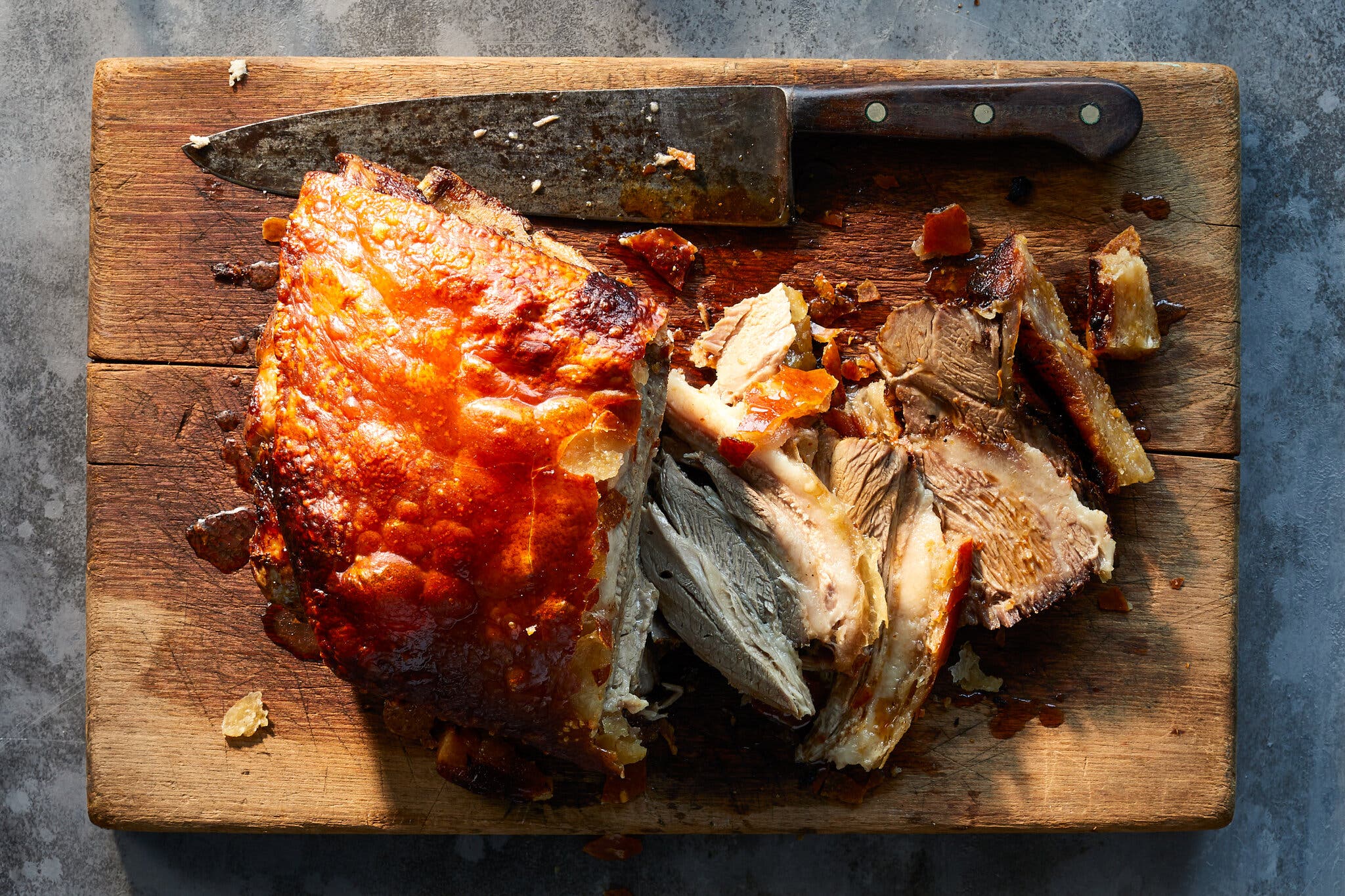 A slab of pernil that has been cut into sits on a cutting board below a large knife.