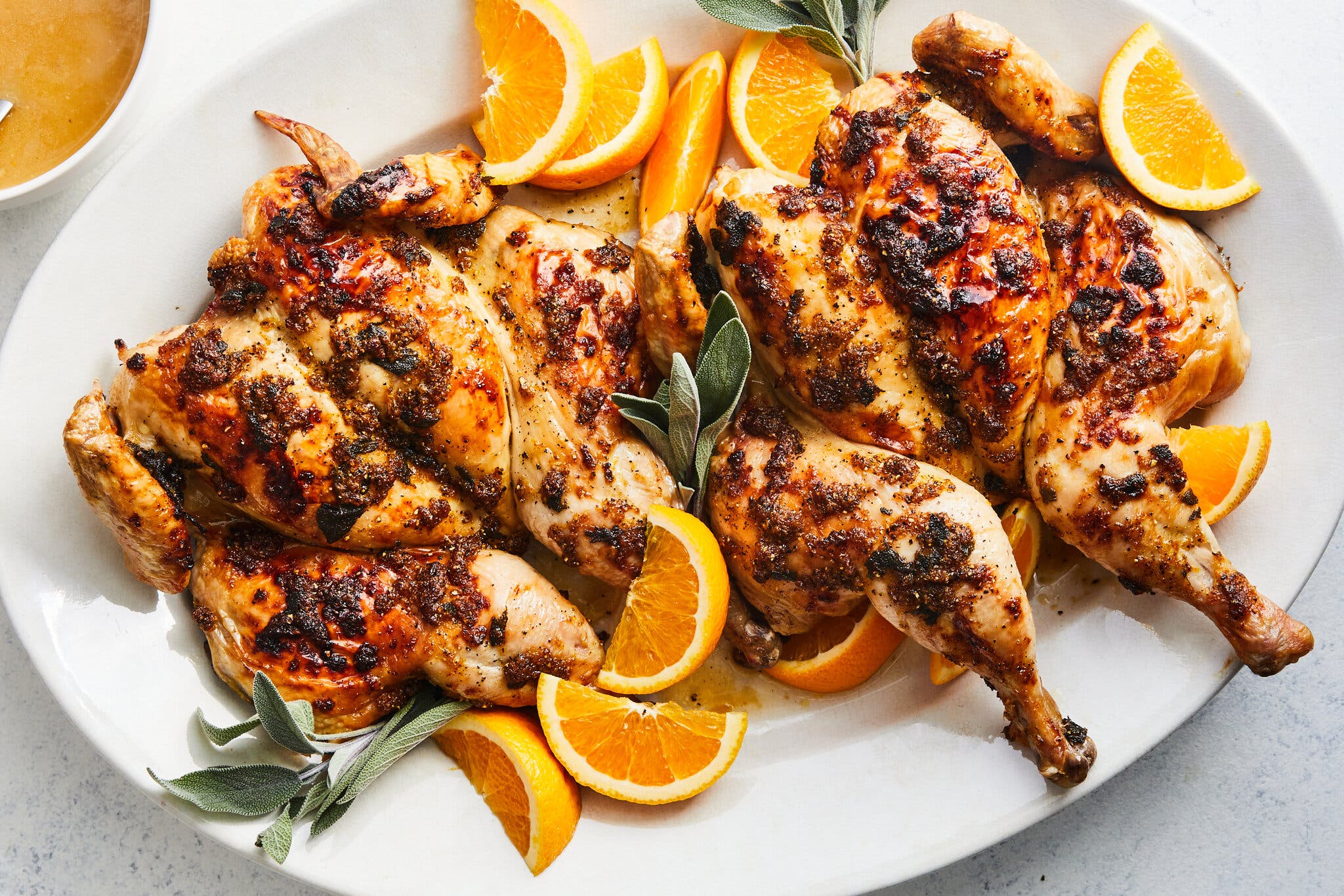 Two roast chickens sit on a large platter surrounded by oranges and bay leaves.