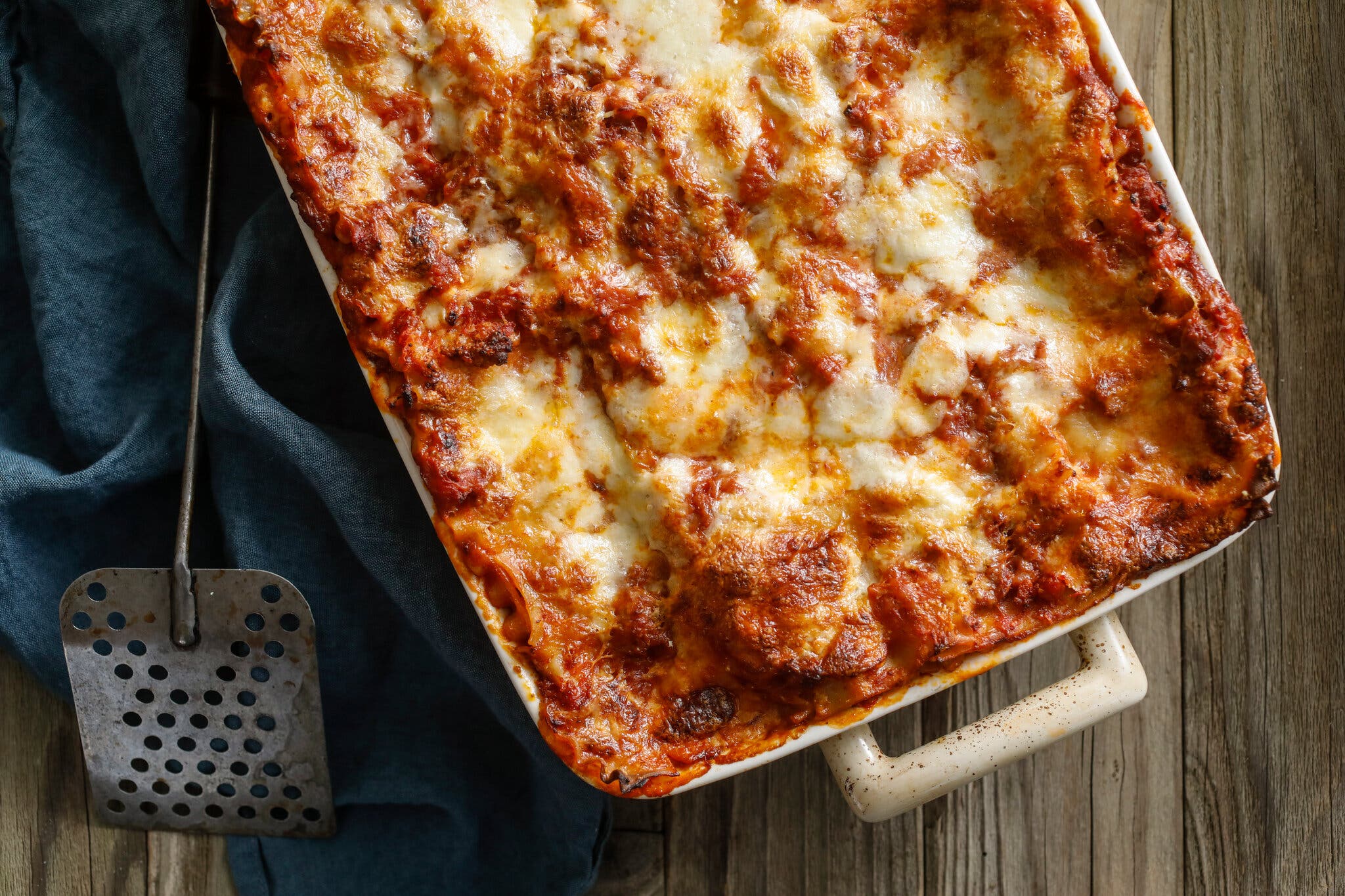 A lasagna in a ceramic dish sits on top of a piece of blue cloth next to a spatula.