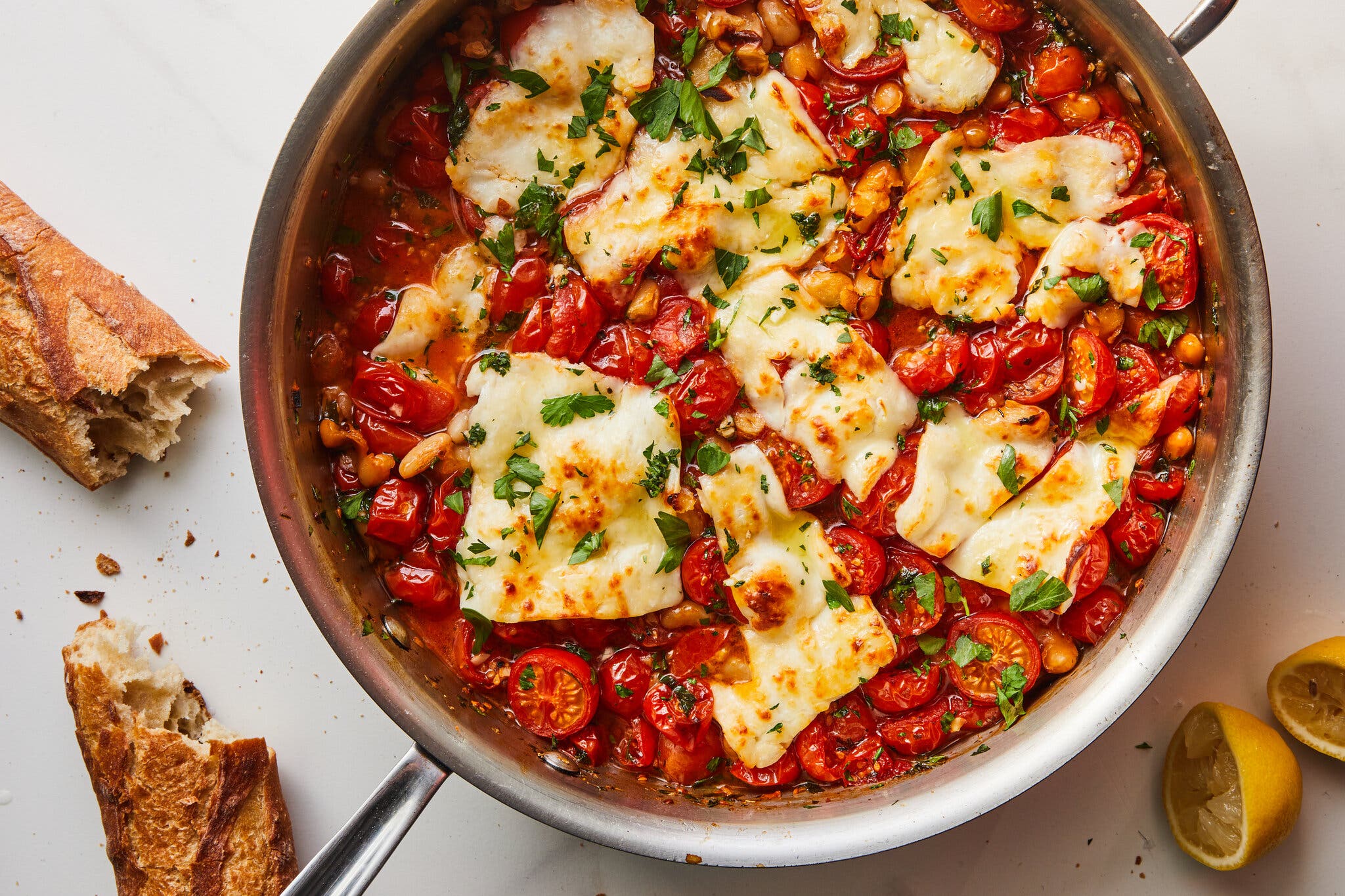 Chunks of halloumi swim in a tomato and white bean stew in a pan. A squeezed lemon and a baguette surround the pan.