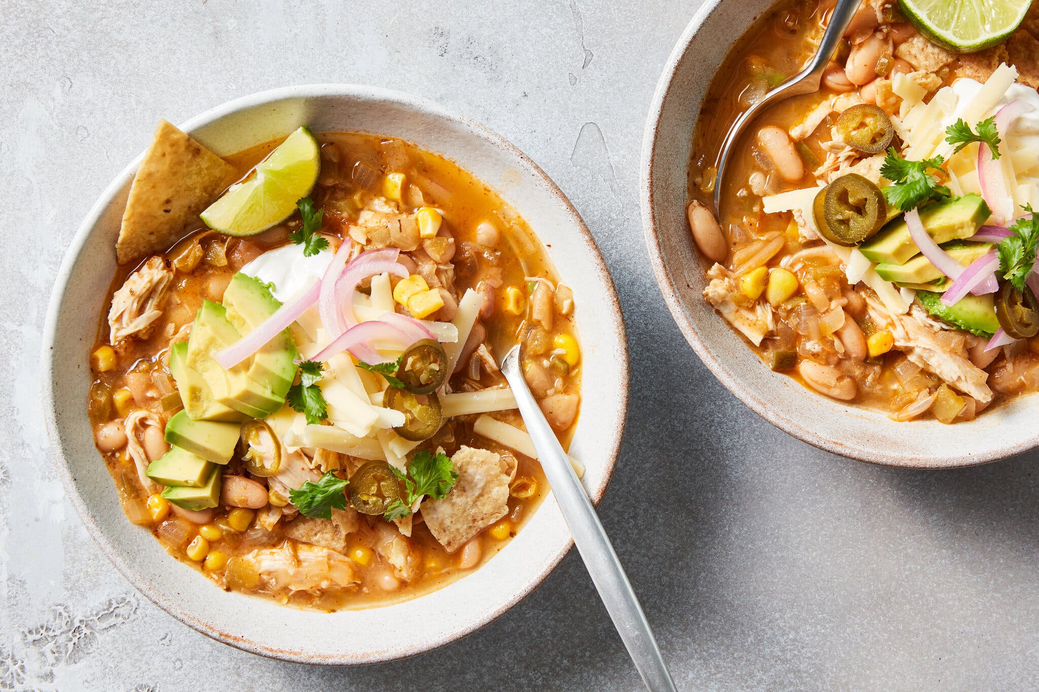 Two bowls of white chicken chili topped with red onions, tortilla chips, avocado, lime and jalapeños.