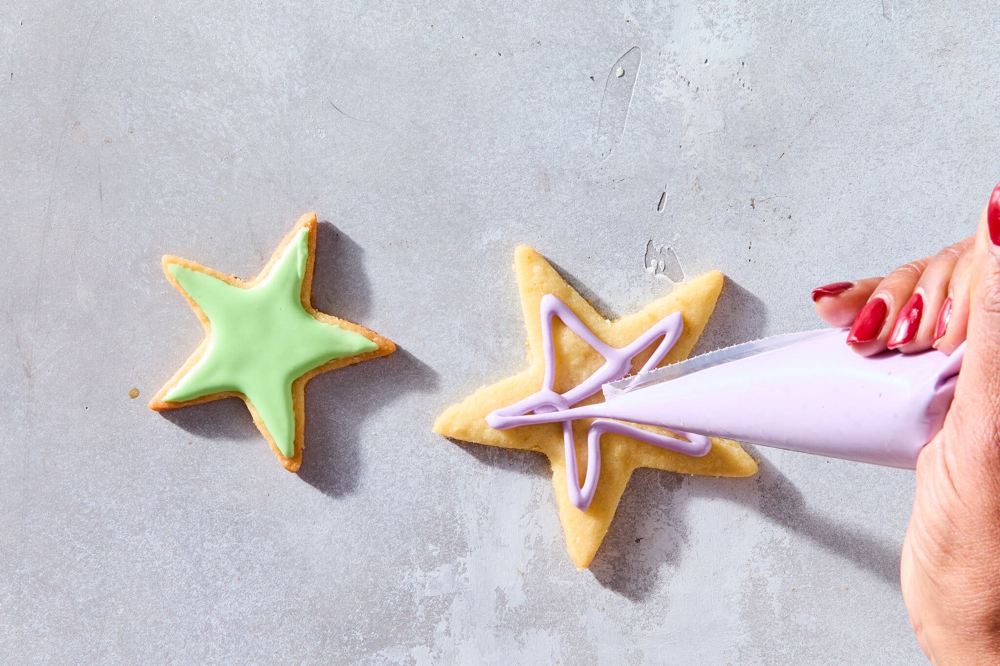 An overhead image of two cookies with colorful icing on top. A hand holding a piping bag hovers over one, the piping still in progress.