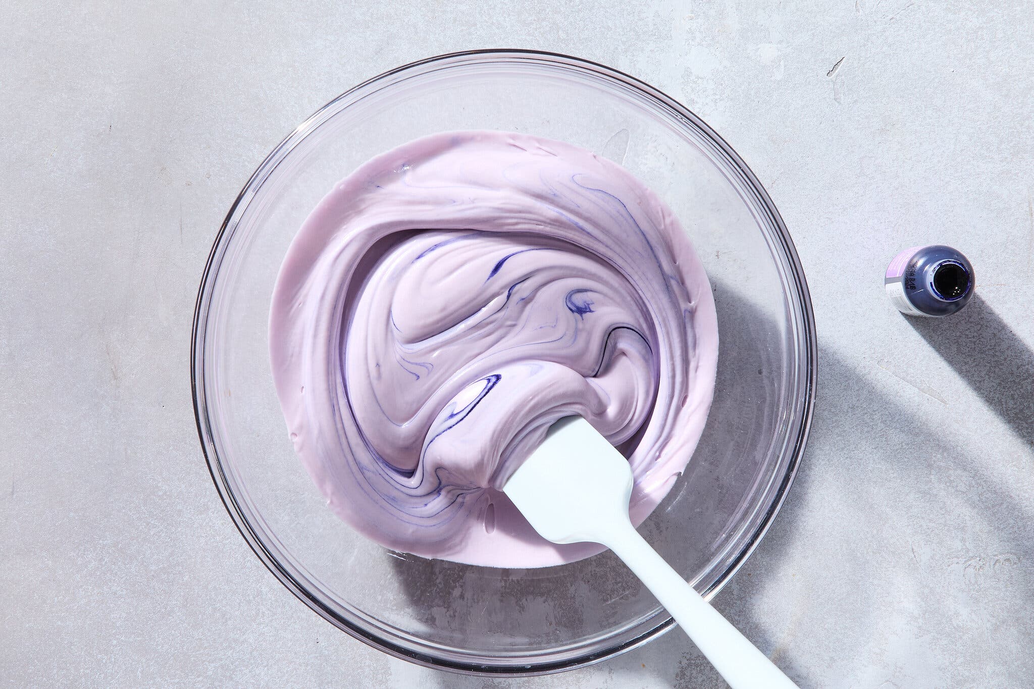 An overhead image of icing in a glass bowl that is being tinted a light purple.
