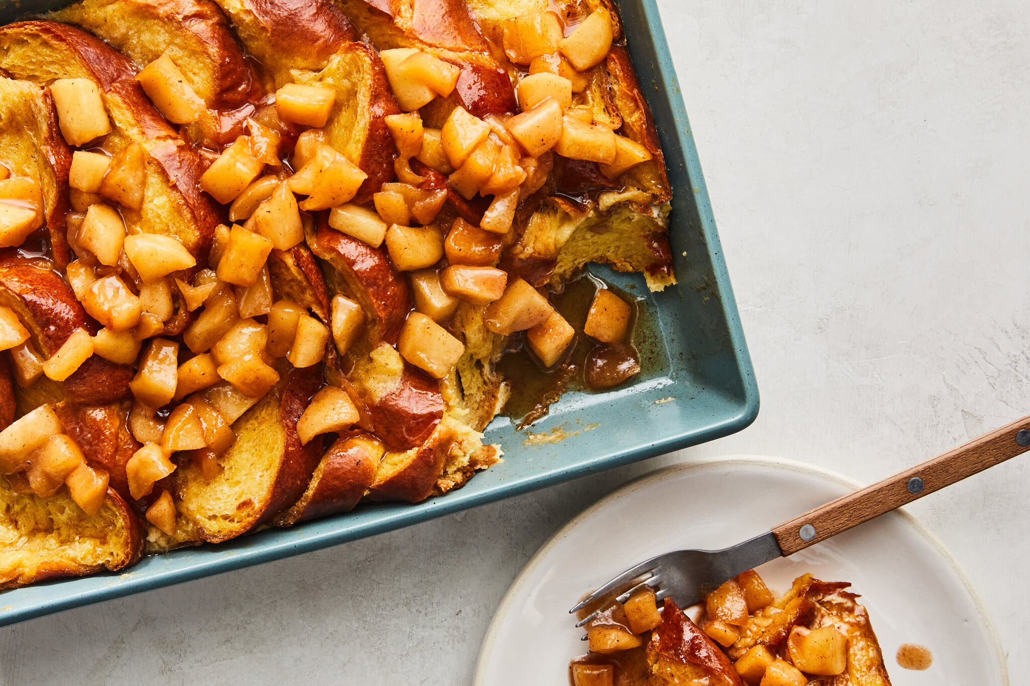 A teal baking dish holds golden-brown French toast topped with shiny, diced apples. A white plate in the foreground contains a serving with a fork.