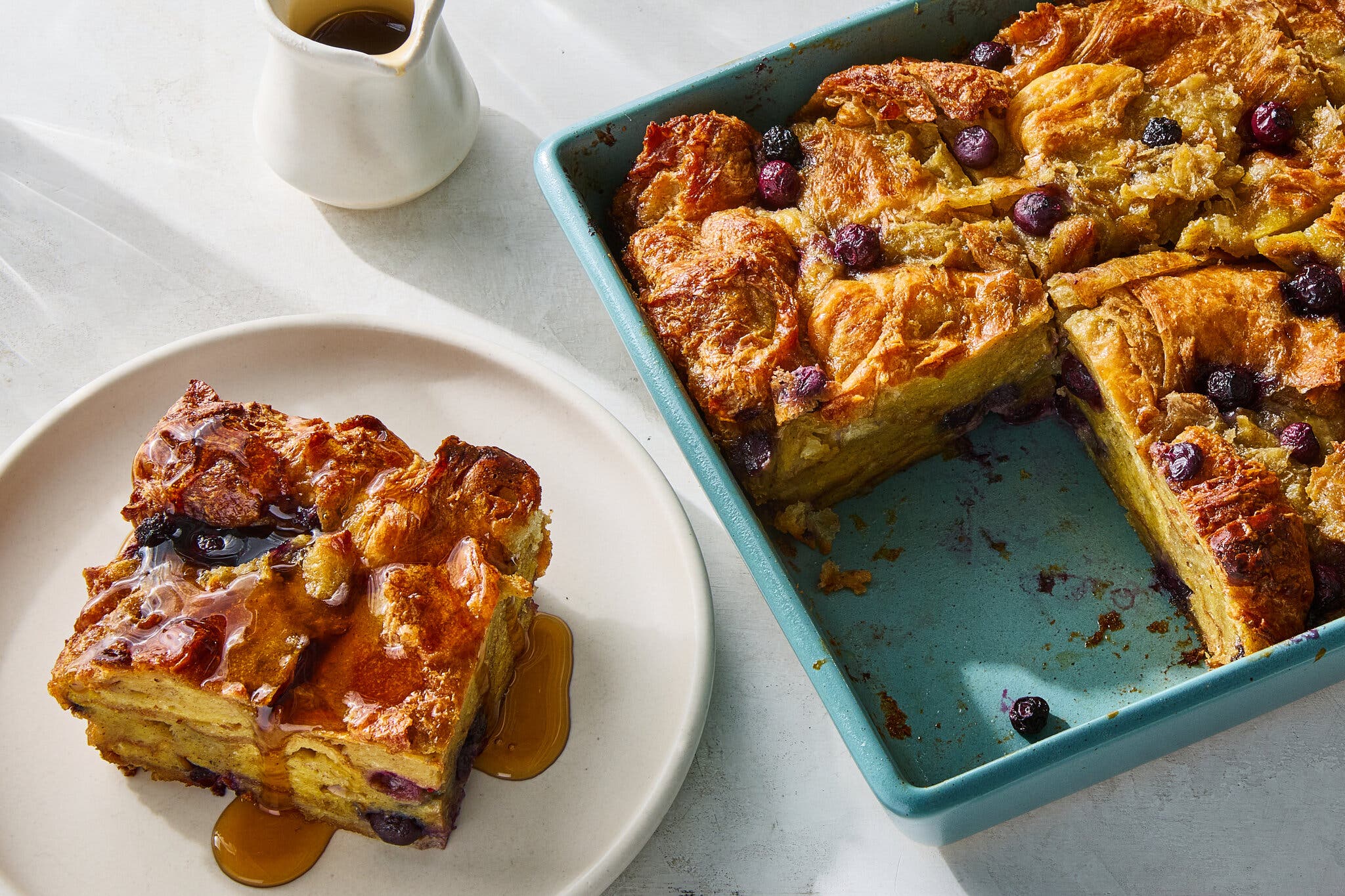 A golden-brown baked French toast casserole with blueberries, one slice served on a plate drizzled with syrup.