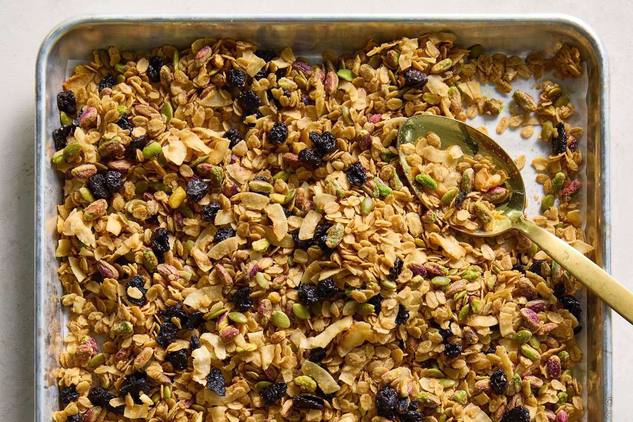 A baking sheet filled with golden granola, green pistachios, dark dried fruit and coconut flakes. A golden spoon sits within the mixture.