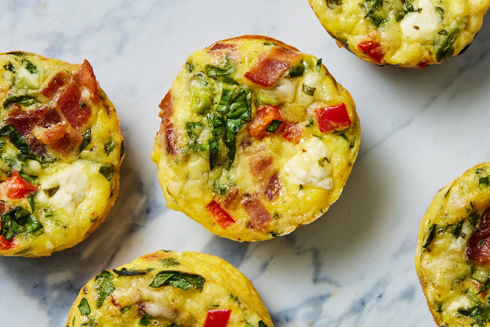 Several egg muffins with green herbs, red bell pepper, white cheese and bacon bits sit on a white marble surface.