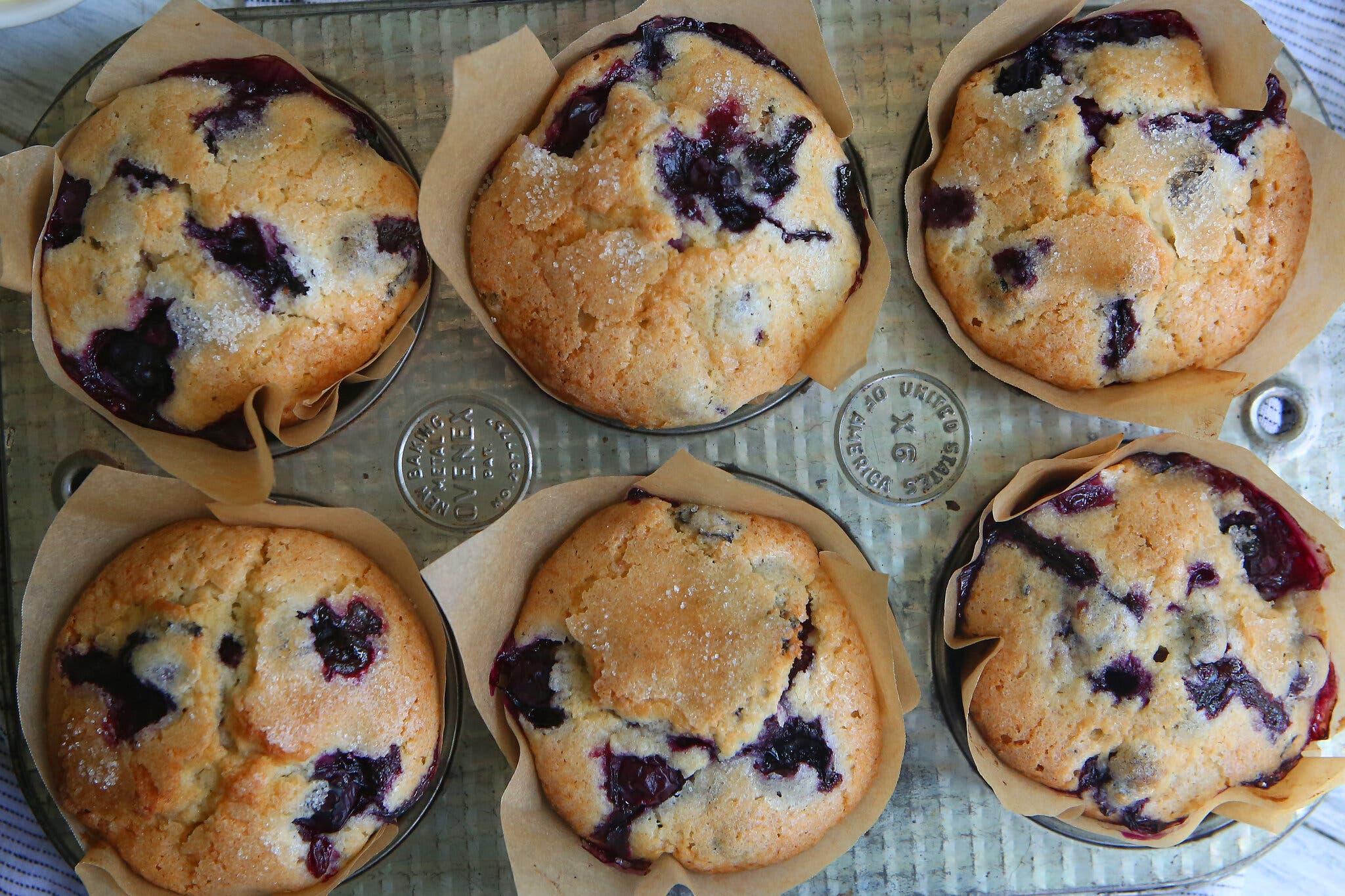 Six golden-brown blueberry muffins with light brown paper liners sit in a metal baking pan.