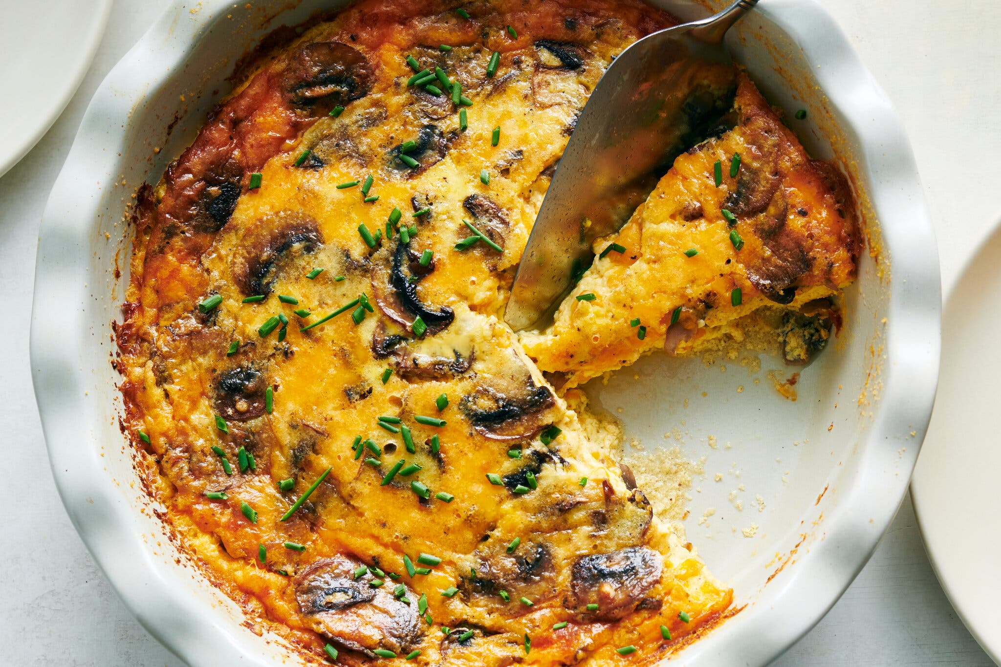 A golden-brown baked quiche filled with mushrooms and topped with green chives. A metal utensil lifts a slice.