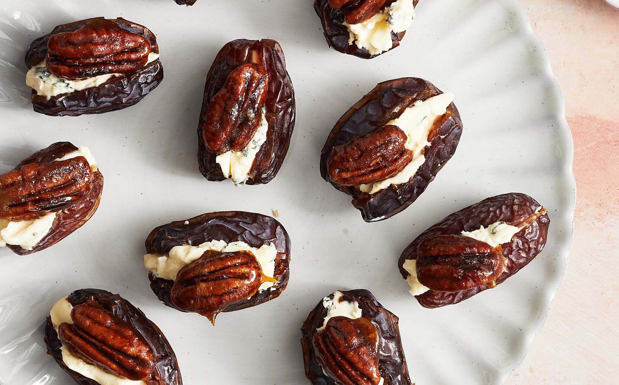 A scalloped white plate with blue-cheese-stuffed dates, each topped with a candied pecan