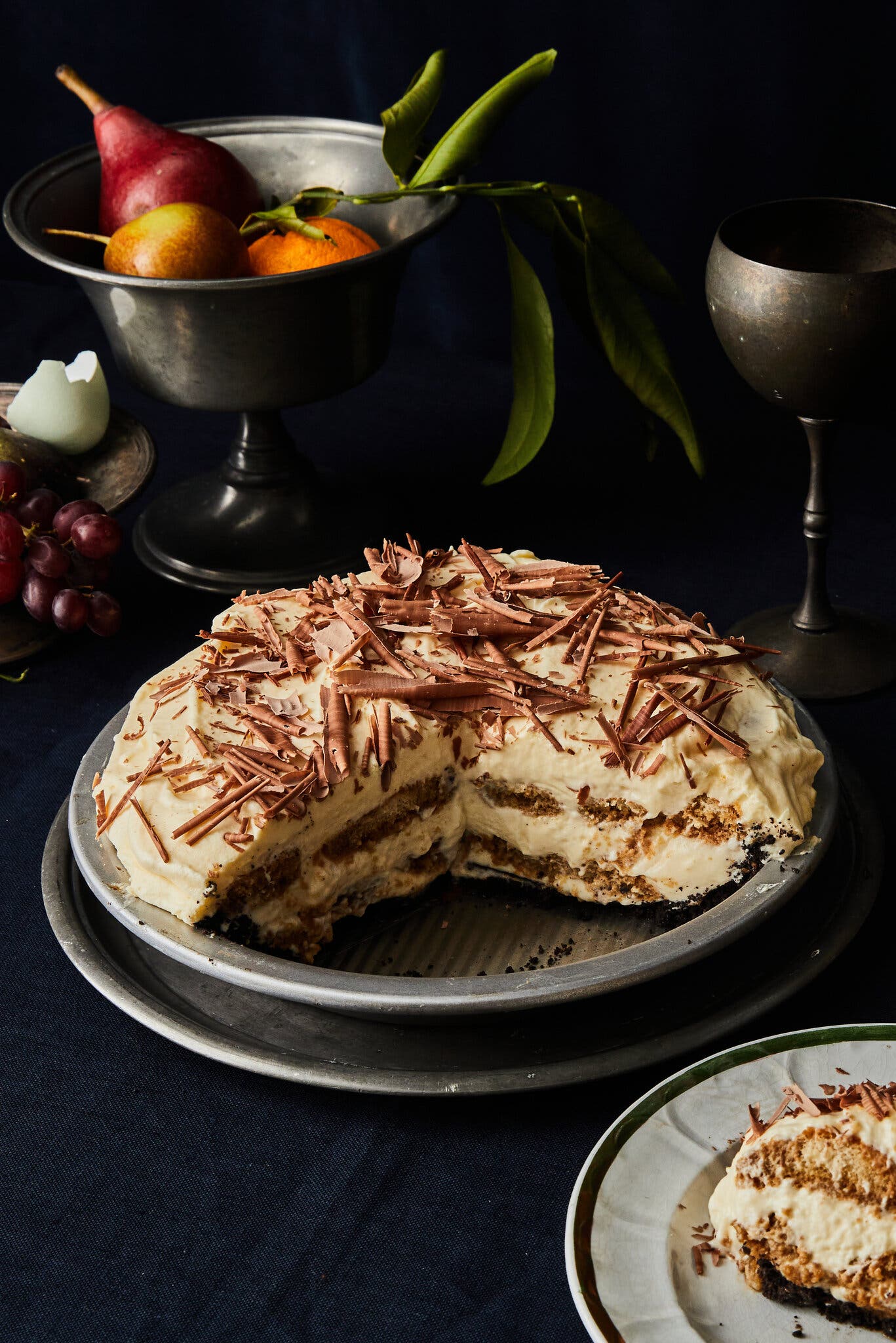 An image of a creamy layered pie topped with chocolate curls. A single piece is taken out. To the back are two silver vessels.
