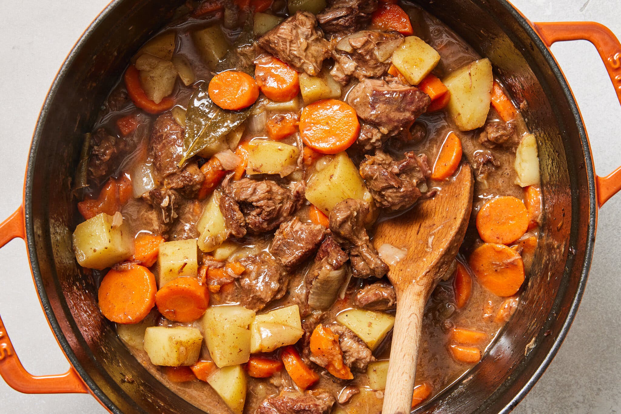 Chunks of beef, circular carrot slices, and cubed potatoes simmering in an orange enameled Dutch oven.