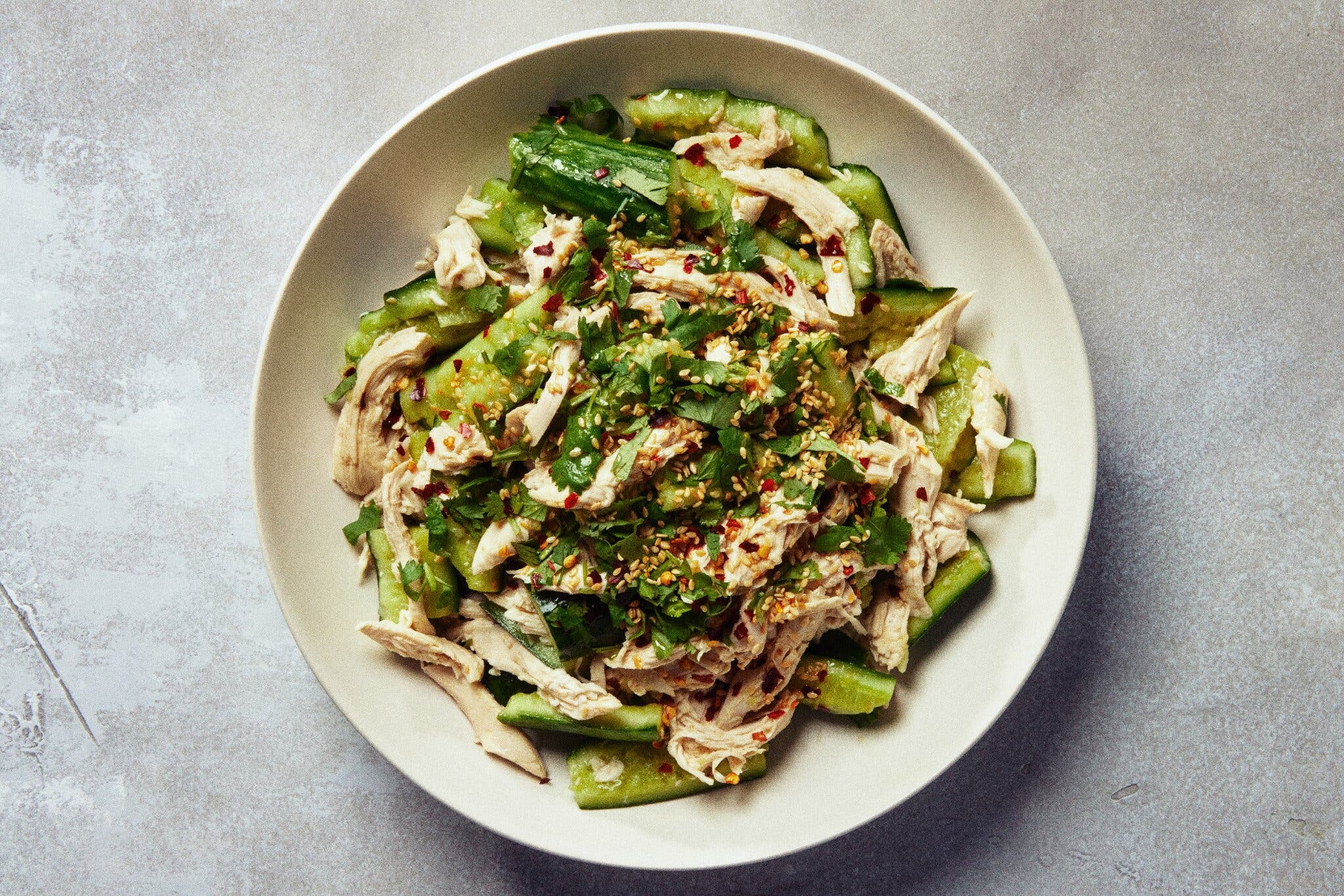 An overhead image of cucumber and chicken in a bowl topped with chile flakes and sesame seeds.